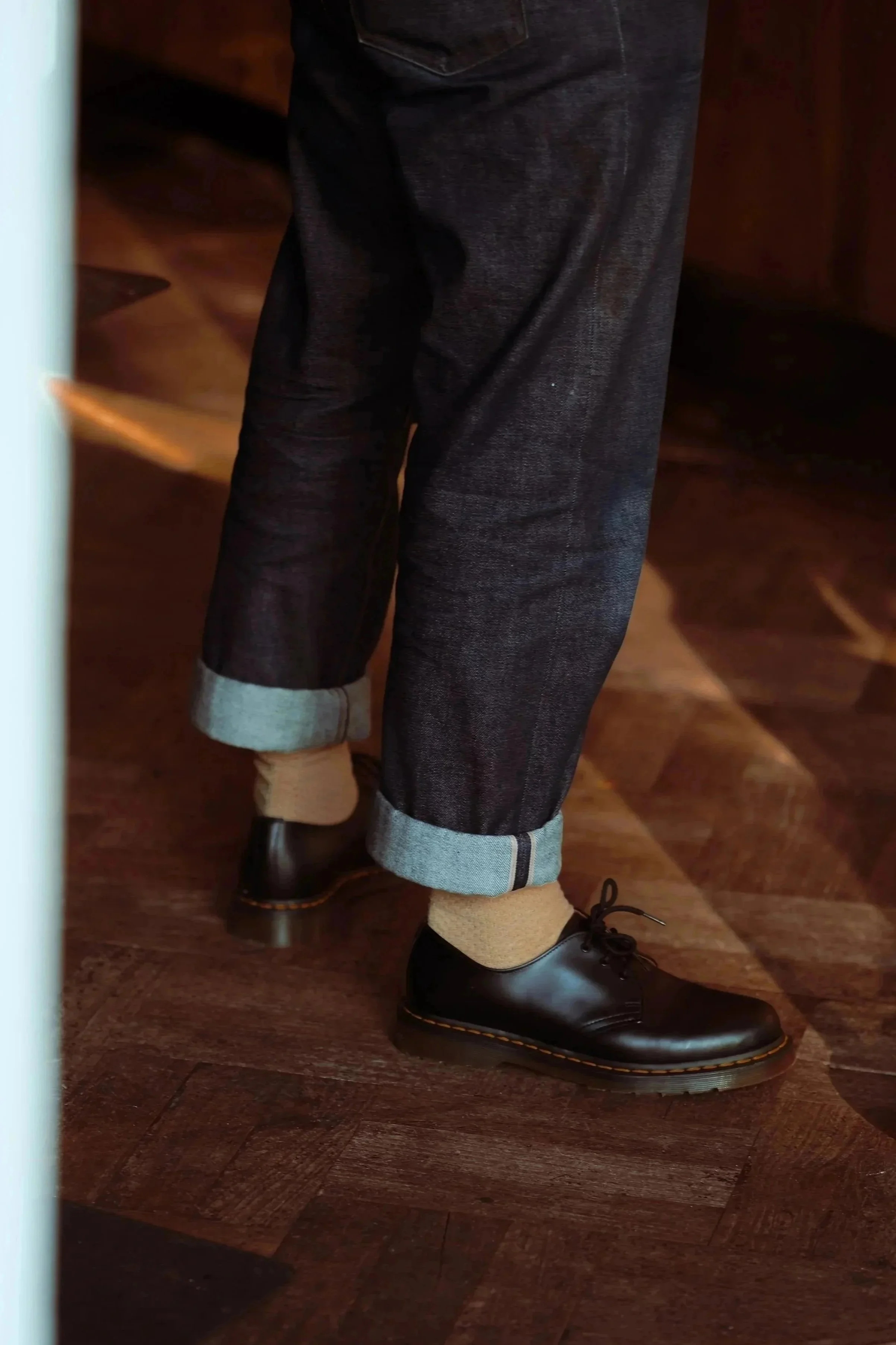 Close-up of a person's black leather shoes, beige socks, dark denim jeans with rolled cuffs, standing on a wooden floor.