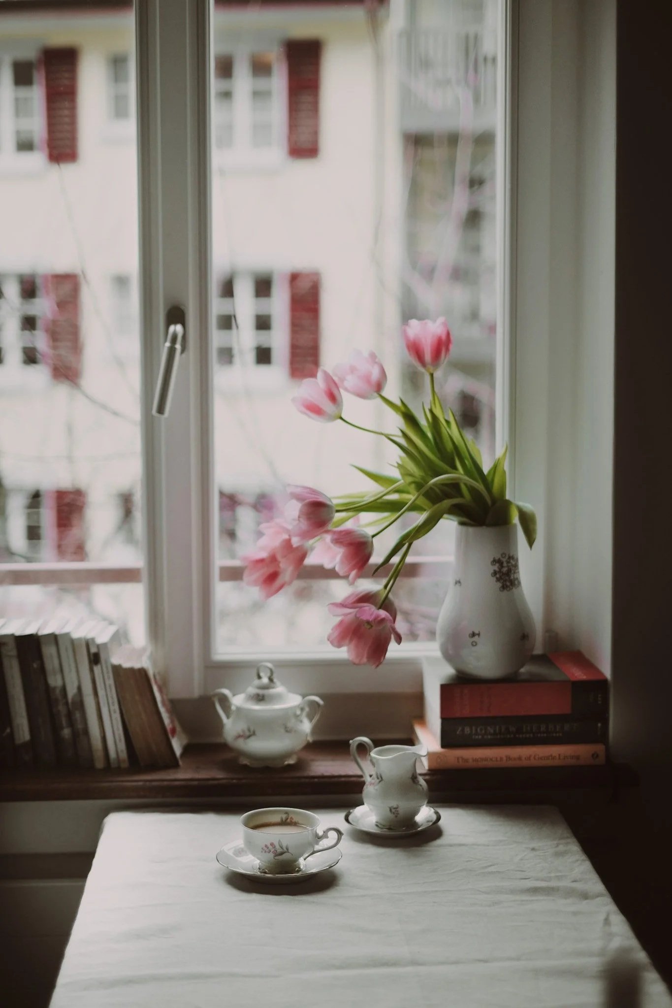 Pink tulips in a white vase on a windowsill, with a teapot, cup, and creamer on a table in front. Books are placed on either side of the window.