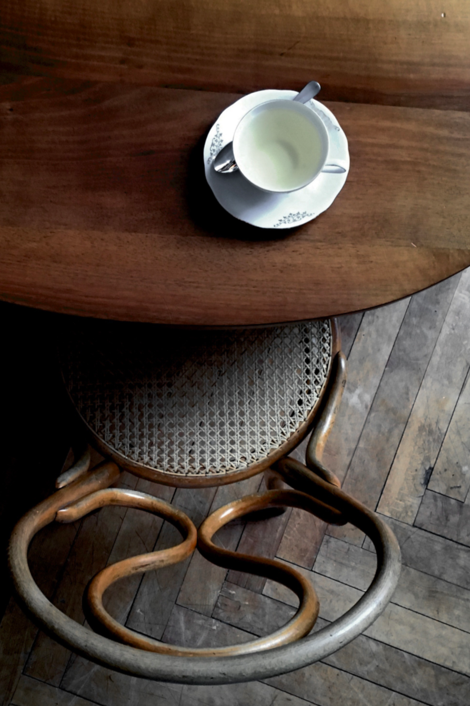 Top-down view of a white cup of coffee or tea with a metallic spoon on a saucer on a wooden table next to a woven cane chair with curved wooden arms. The table has a warm brown finish, and the floor is made of wooden planks.