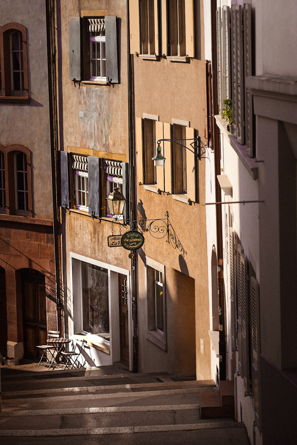 Colorful European street with pastel buildings, open shutters, outdoor seating, and hanging lanterns, in warm sunlight.