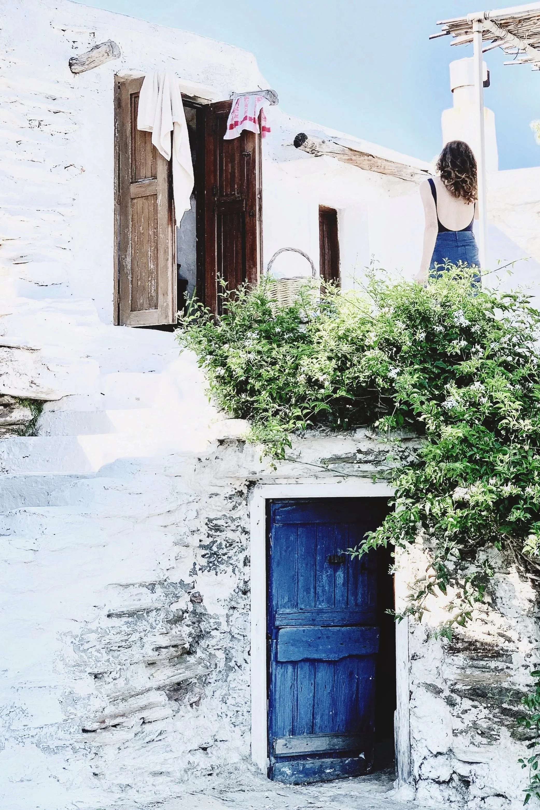 A woman with curly hair wearing a blue dress standing outside of a white stone house with a blue wooden door, greenery, and a laundry line with towels.