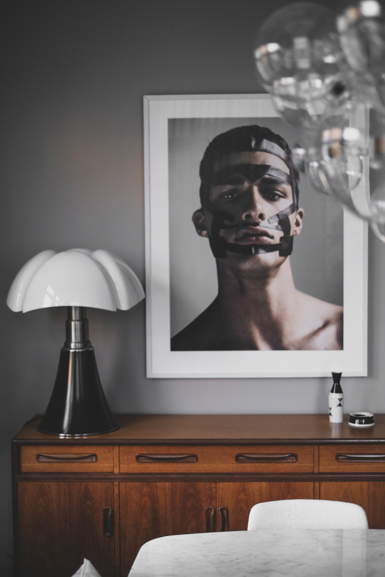 A modern interior with a large framed black and white portrait of a young man on the wall, a wooden sideboard, a white and black table lamp, and a white chair, with glass pendant lights hanging from the ceiling.