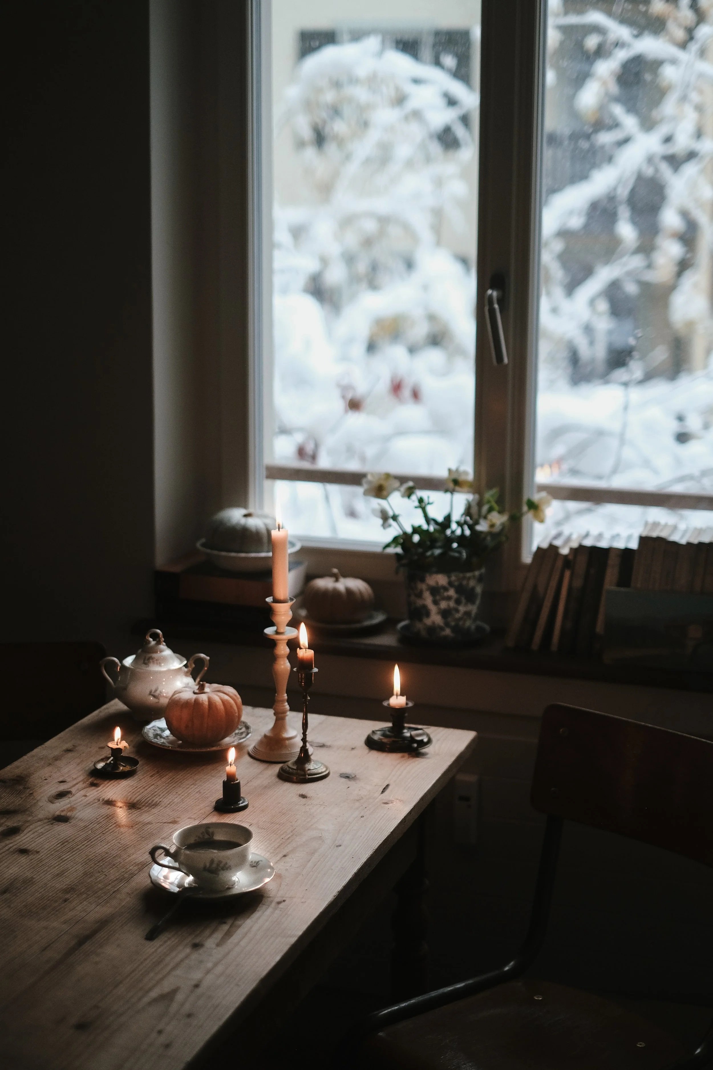 A cozy indoor scene with a wooden table set with a teacup, saucer, and lit candles, looking out through a window at snowy trees outside.