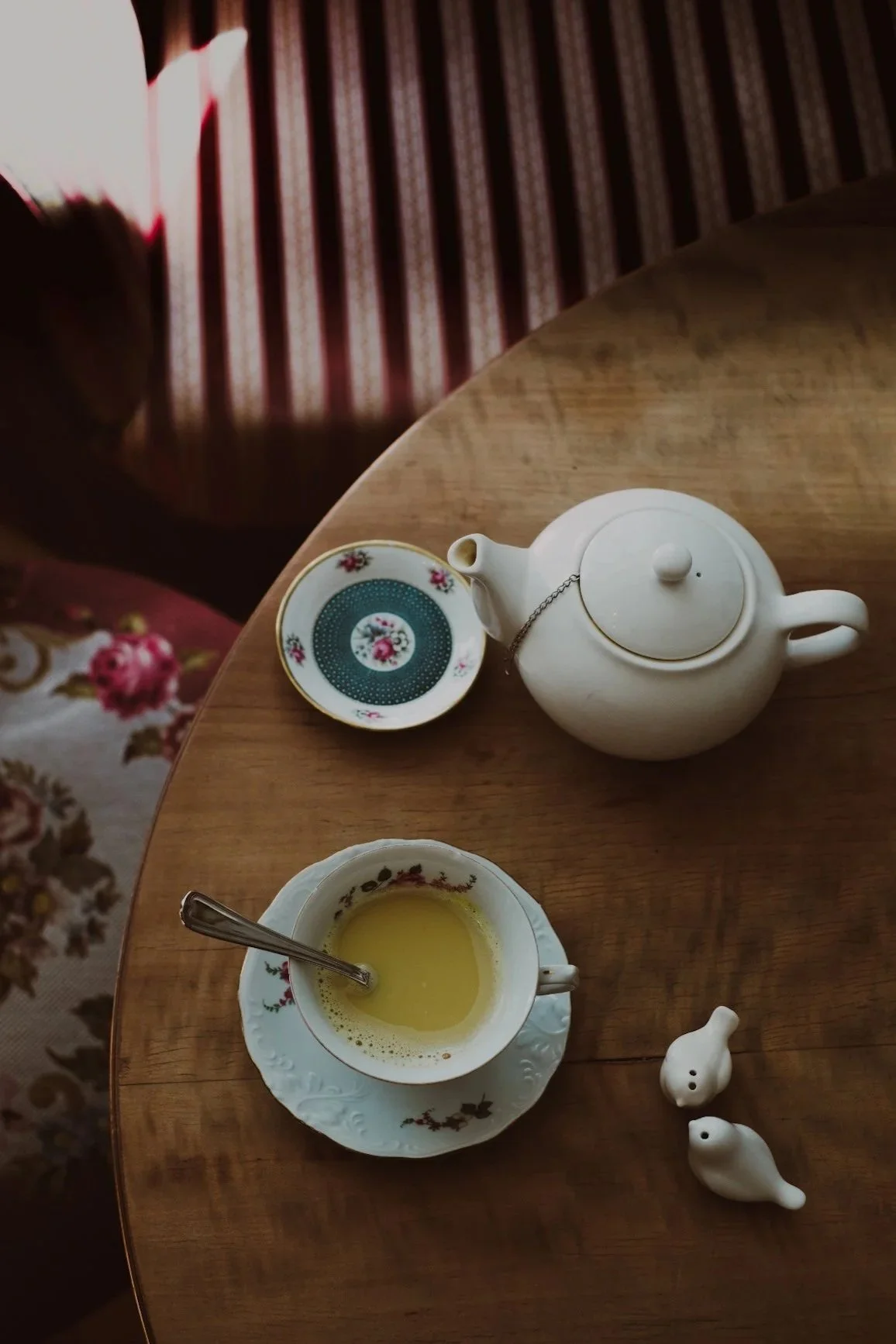 A teapot, a teacup with yellow liquid and a spoon, a small dish with a floral pattern, and two small salt and pepper shakers shaped like birds on a wooden table.