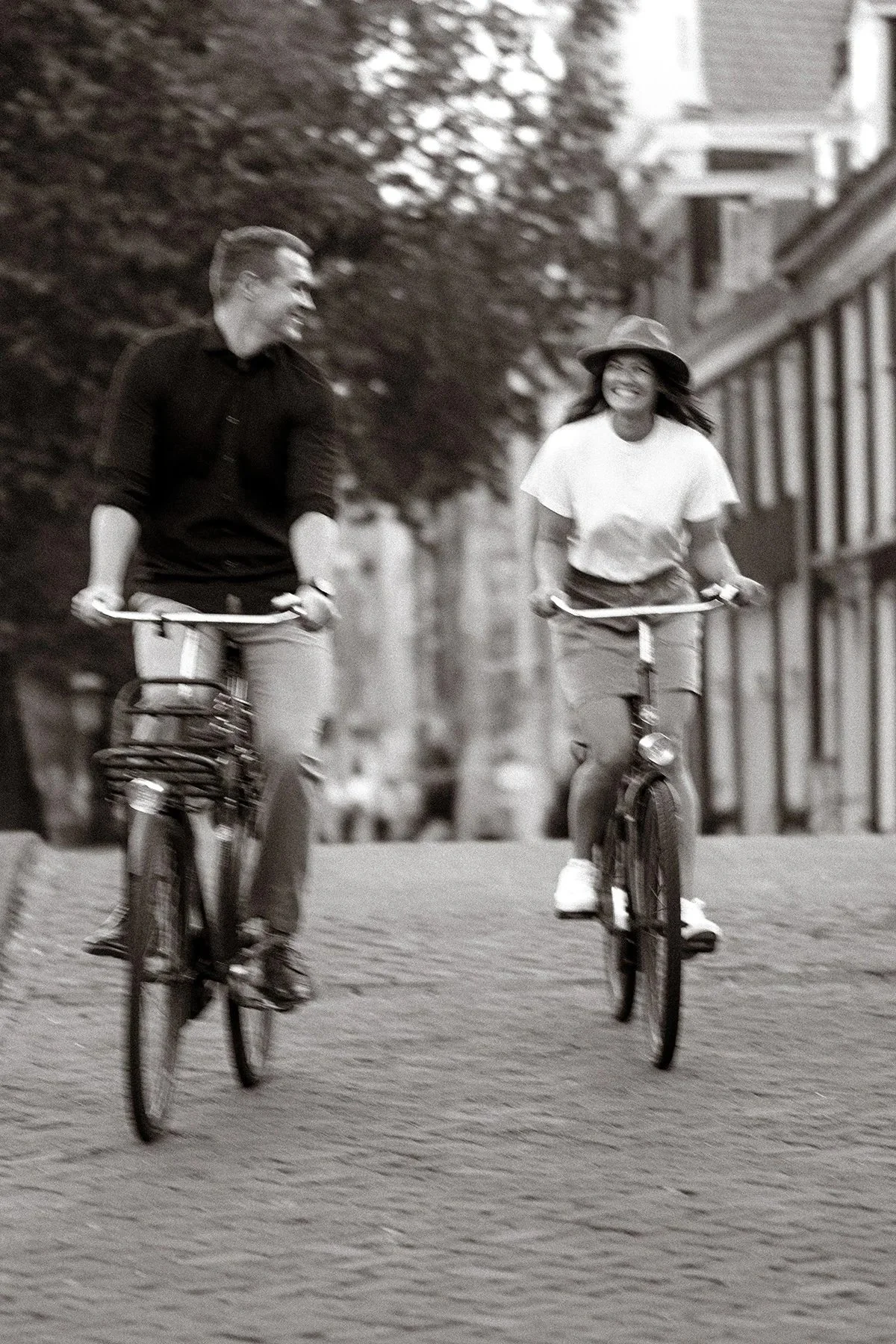 Two people riding bicycles and smiling in conversation on a city street with trees and buildings in the background.