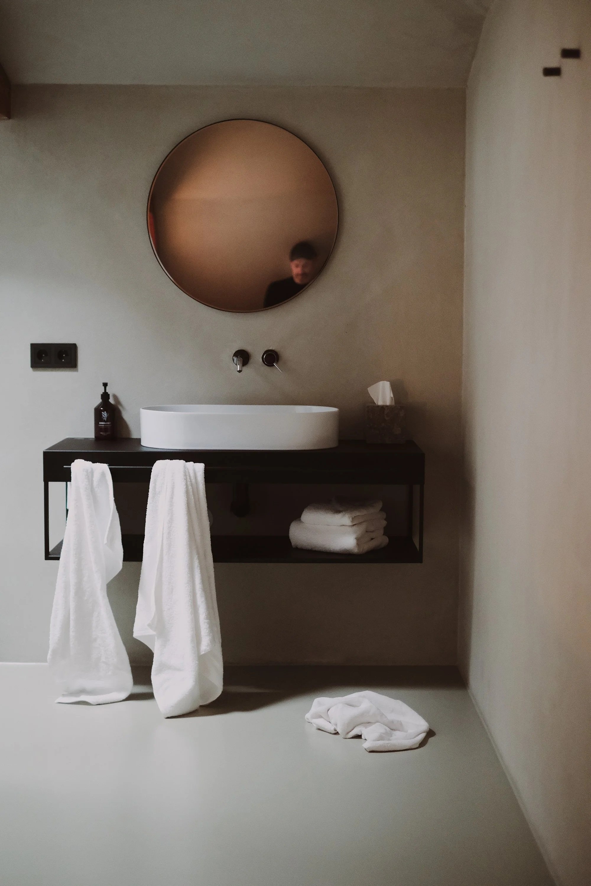 Minimalist bathroom with a round mirror, black vanity with a vessel sink, two hanging white towels, folded towels on a lower shelf, a bottle of soap, tissue box, and a crumpled white cloth on the floor.