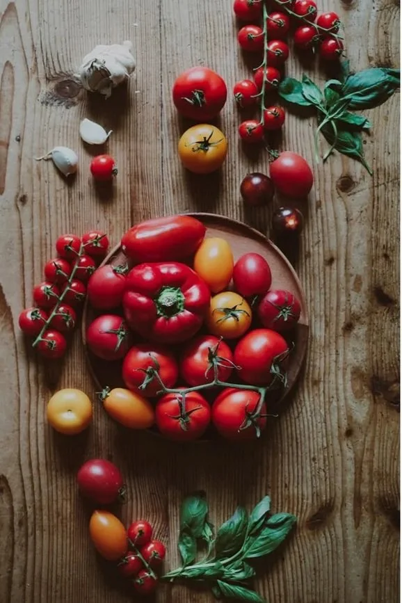 Various colorful tomatoes and garlic on a wooden table, with a basket of cherry tomatoes and a pink flower in a vase nearby.