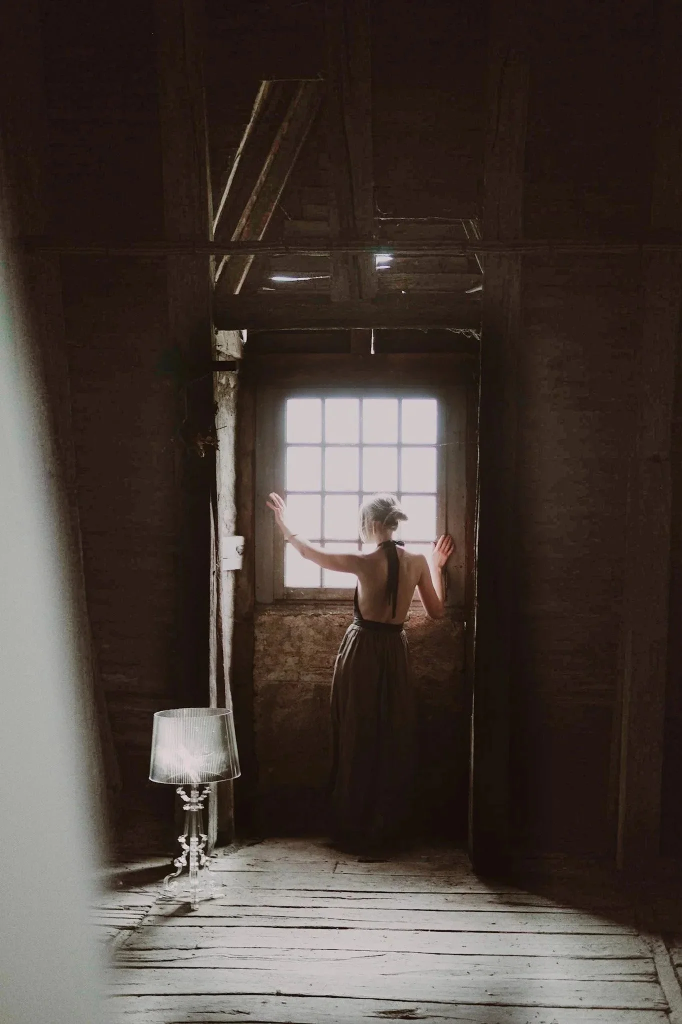 A woman with blonde hair in a bun, wearing a backless dress, stands in a rustic room with wooden walls and floor, facing a window, with her hands touching the wall on either side of the window, and a small table with a lamp beside her.