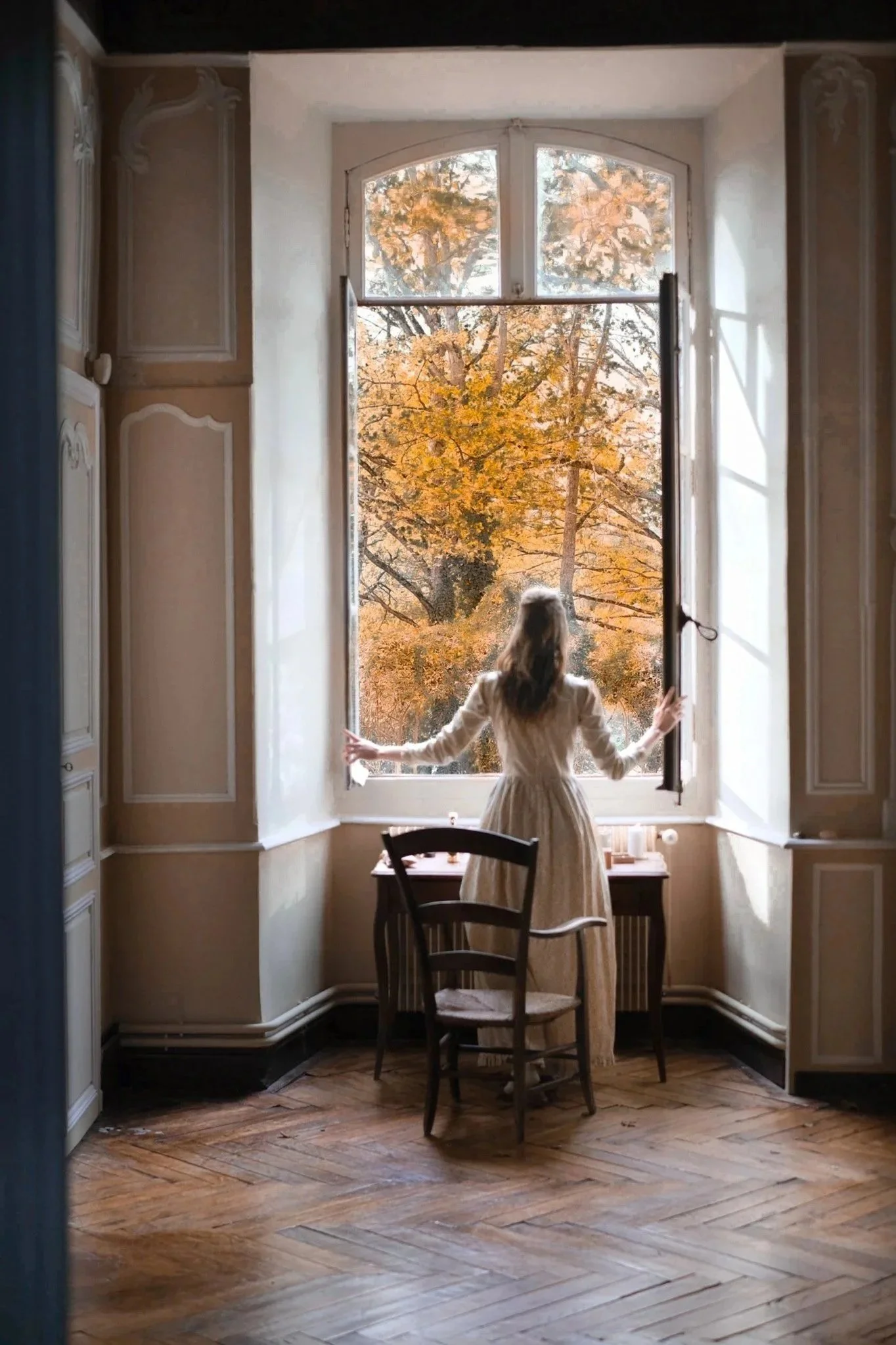 A woman in a cream-colored dress stands at an open window looking outside at autumn trees with orange leaves.