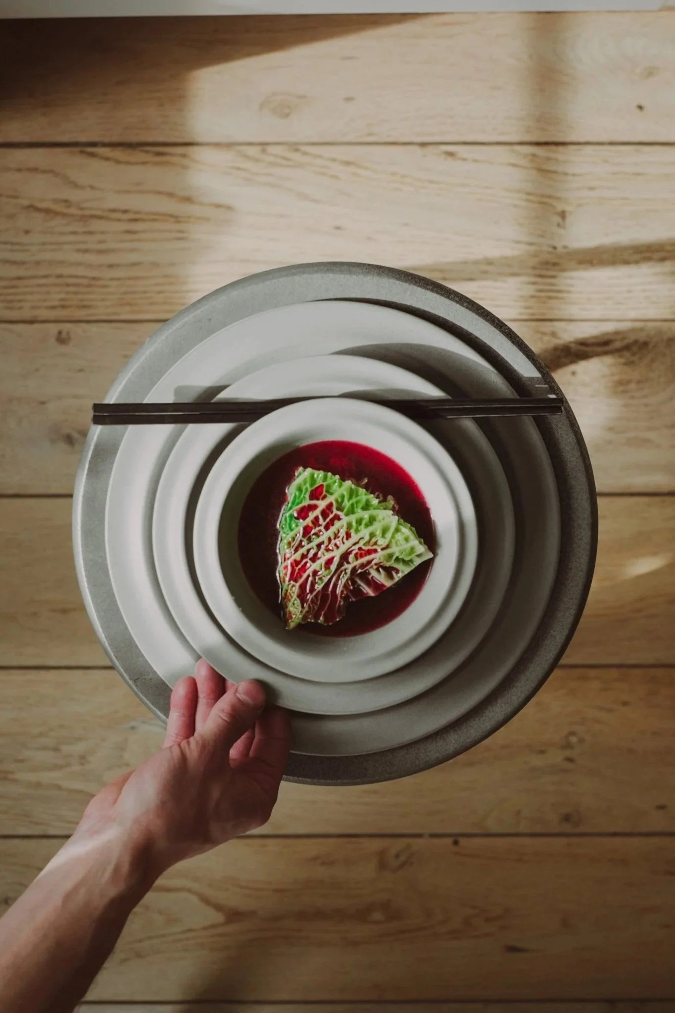 Hand holding a small plate with a piece of salad and a dressing on it, on layered round plates with chopsticks resting on top, wooden floor background.