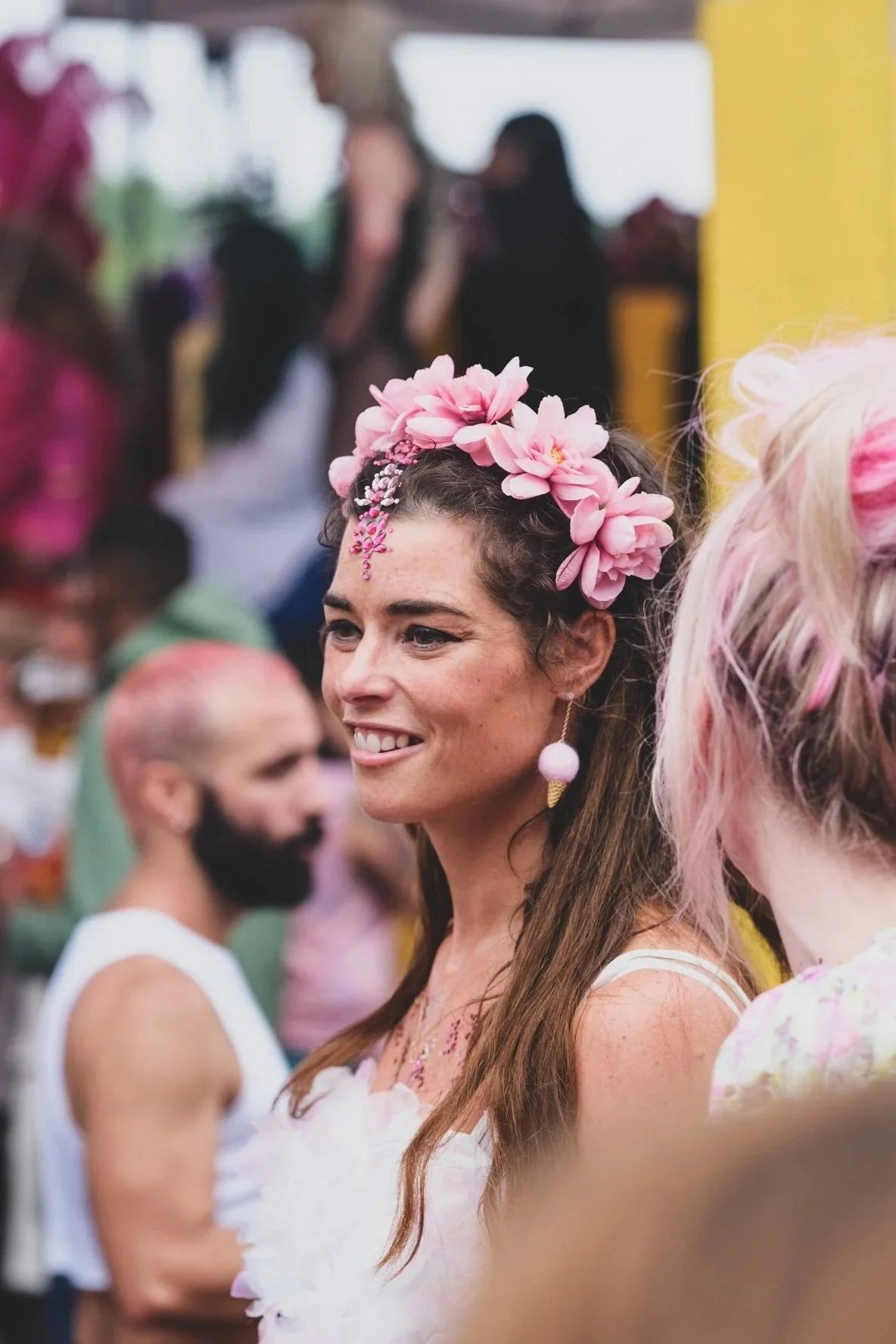 A woman with a smile, wearing a pink floral headpiece and an earring, at a colorful outdoor event with blurred people in the background.