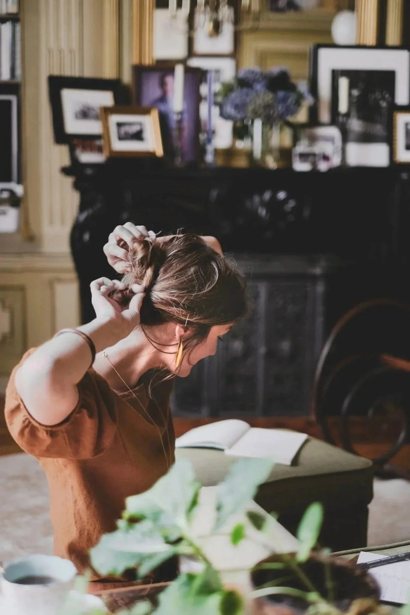 A woman tying her hair in a cozy, well-decorated living room with framed photos, a vase with purple flowers, and a fireplace.