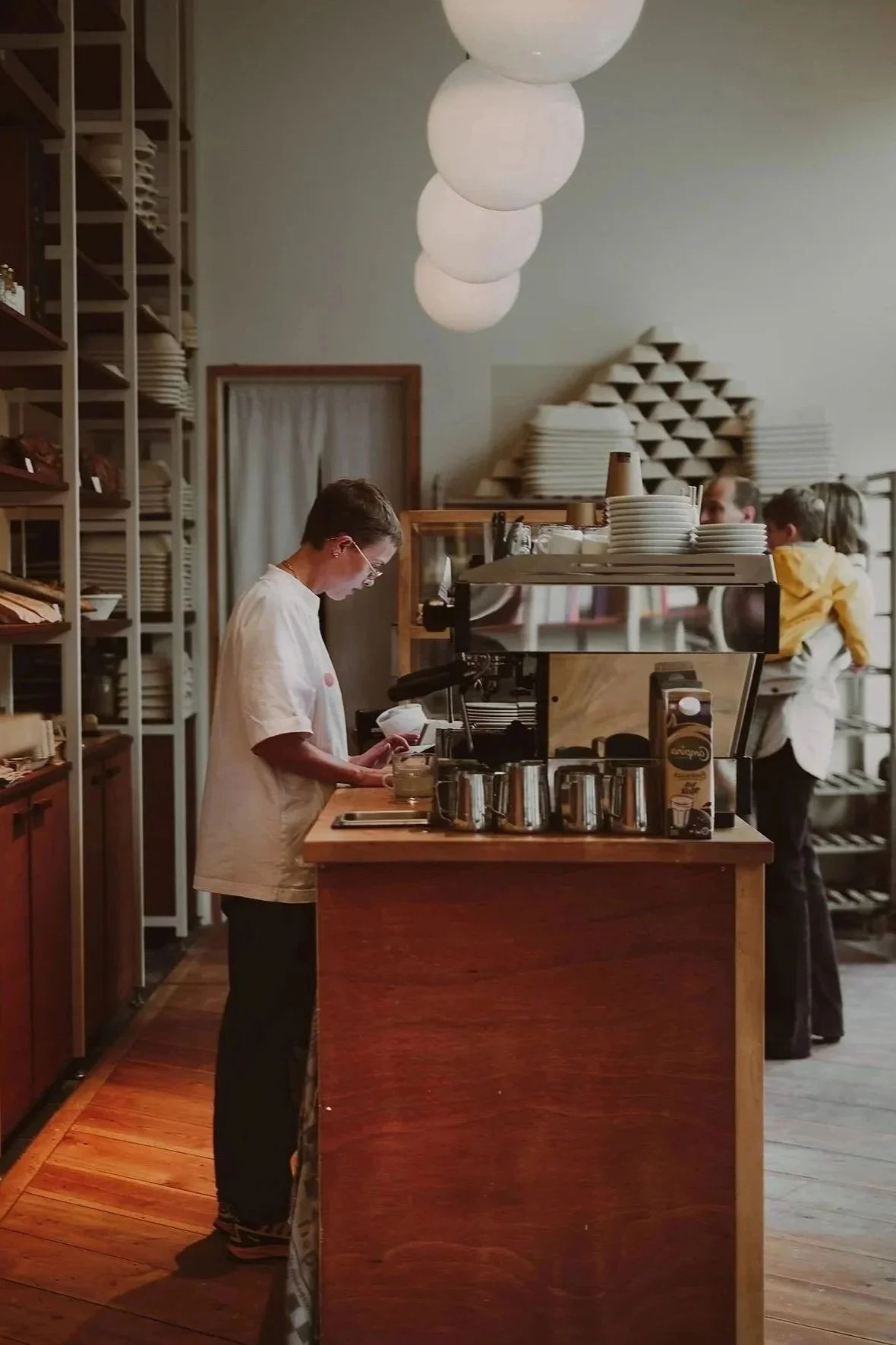 A barista working behind a wooden counter in a coffee shop, with shelves of plates and cups on the wall and large white hanging lights overhead.