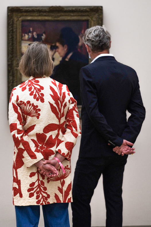 Two people, a woman and a man, observing a painting in an art gallery. The woman is wearing a white coat with large red floral patterns, and the man is dressed in a dark suit. They are standing close together, facing the artwork.