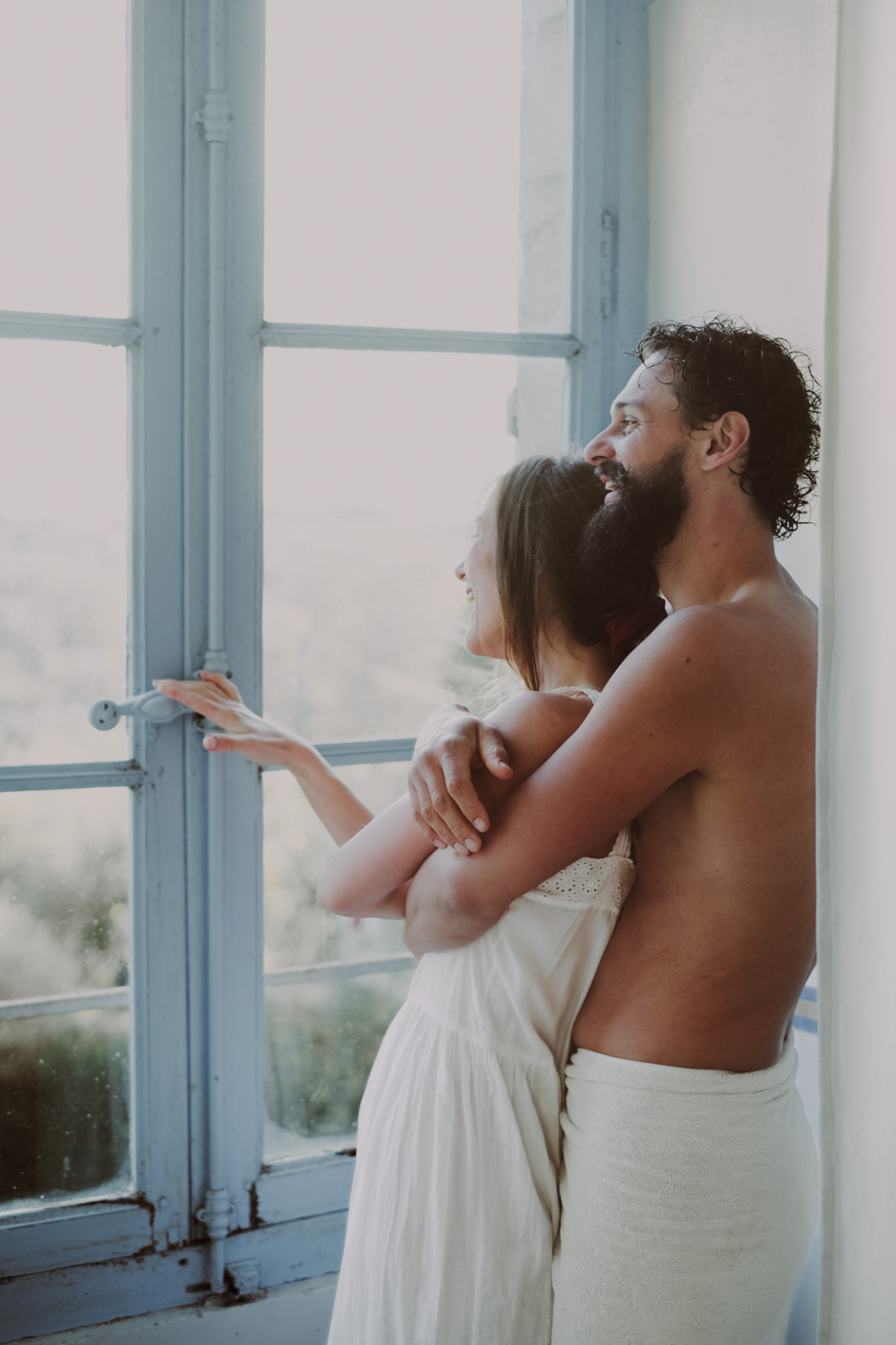 A man and woman, both in sleepwear, share intimate moments in a bright, cozy home. In one scene, they are standing close, smiling and touching each other's faces. In the other, they are embracing by a large window with natural light streaming in.