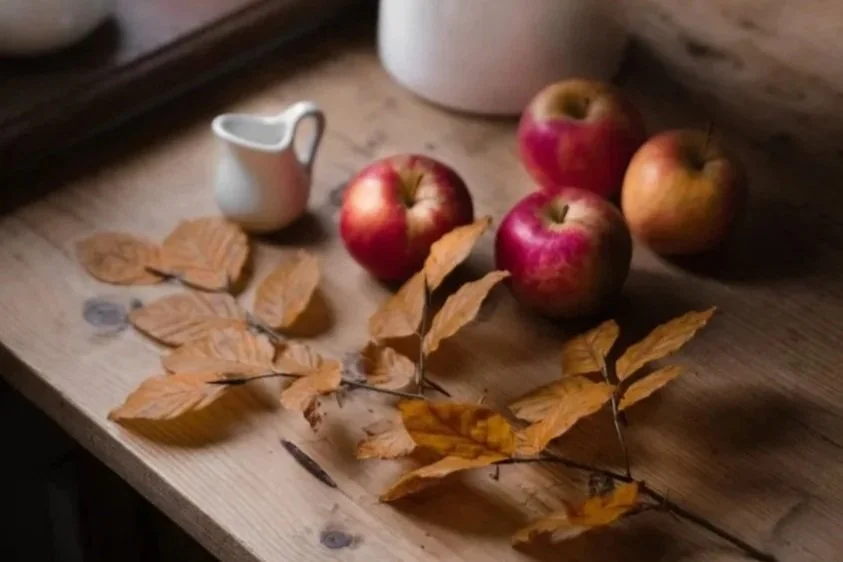 Two photos of a wooden table nearby a window. The first photo shows three apples, a small white creamer, and dried autumn leaves. The second photo shows a bowl of apples, a teacup with saucer, a white pitcher, and a stack of books.