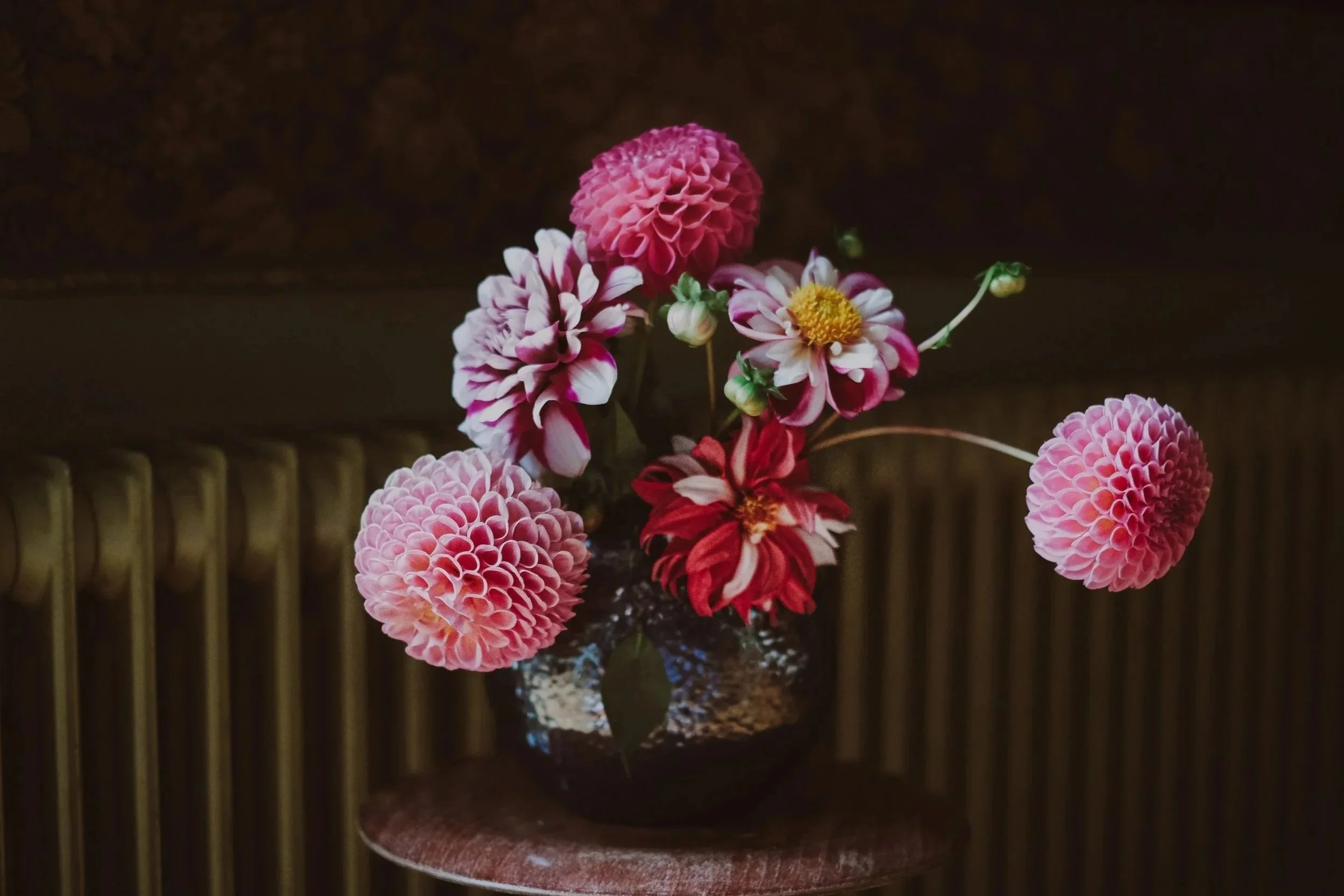 A dark vase with a bouquet of pink, white, and red flowers, including dahlias and other blooms, is placed on a small wooden table against a dark background.