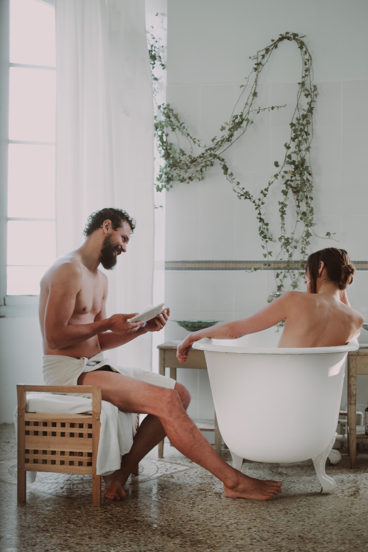 A split image showing a man and a woman in a vintage-style bathroom. The man, with a beard, is sitting on a small wooden stool, reading a book, with a towel around his waist. On the left, the woman is in a bathtub, resting her head on her arm with he