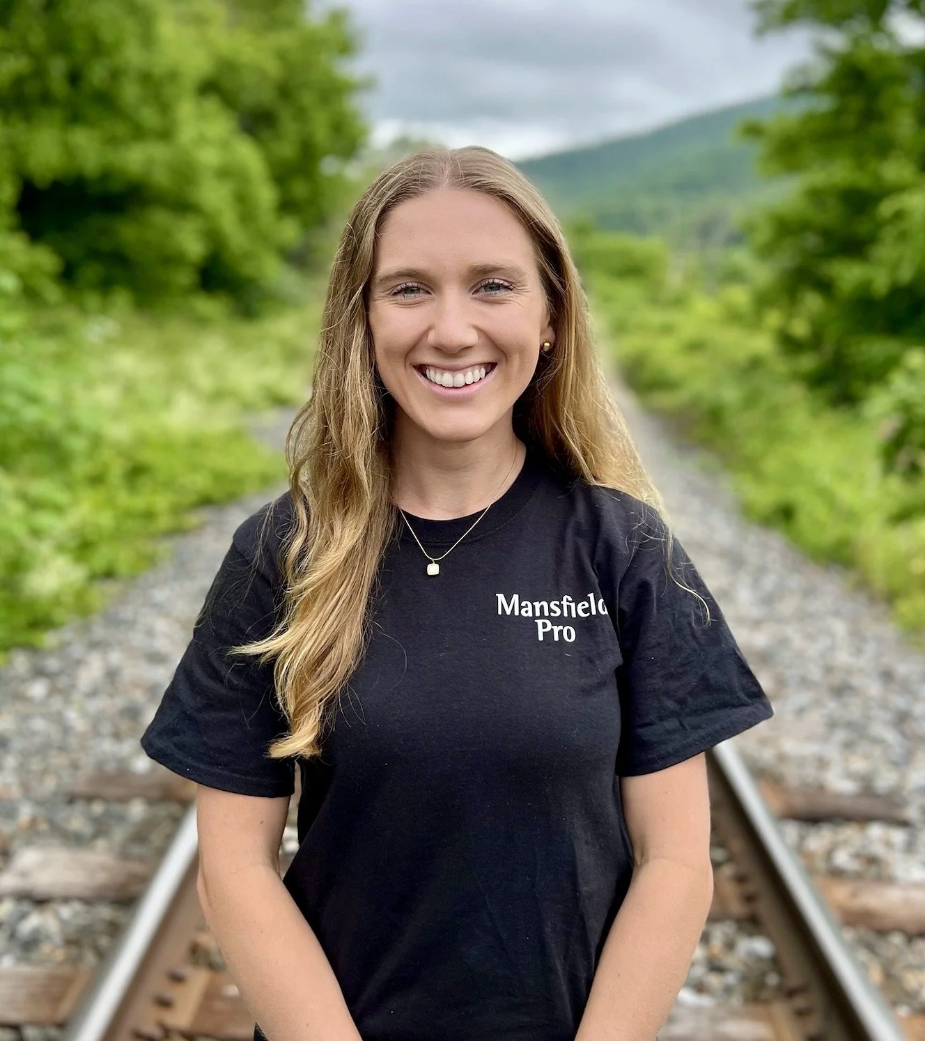 Smiling young woman with long wavy blonde hair standing on railroad tracks in a green natural setting, wearing a black T-shirt with white text that says "Mansfield Pro."