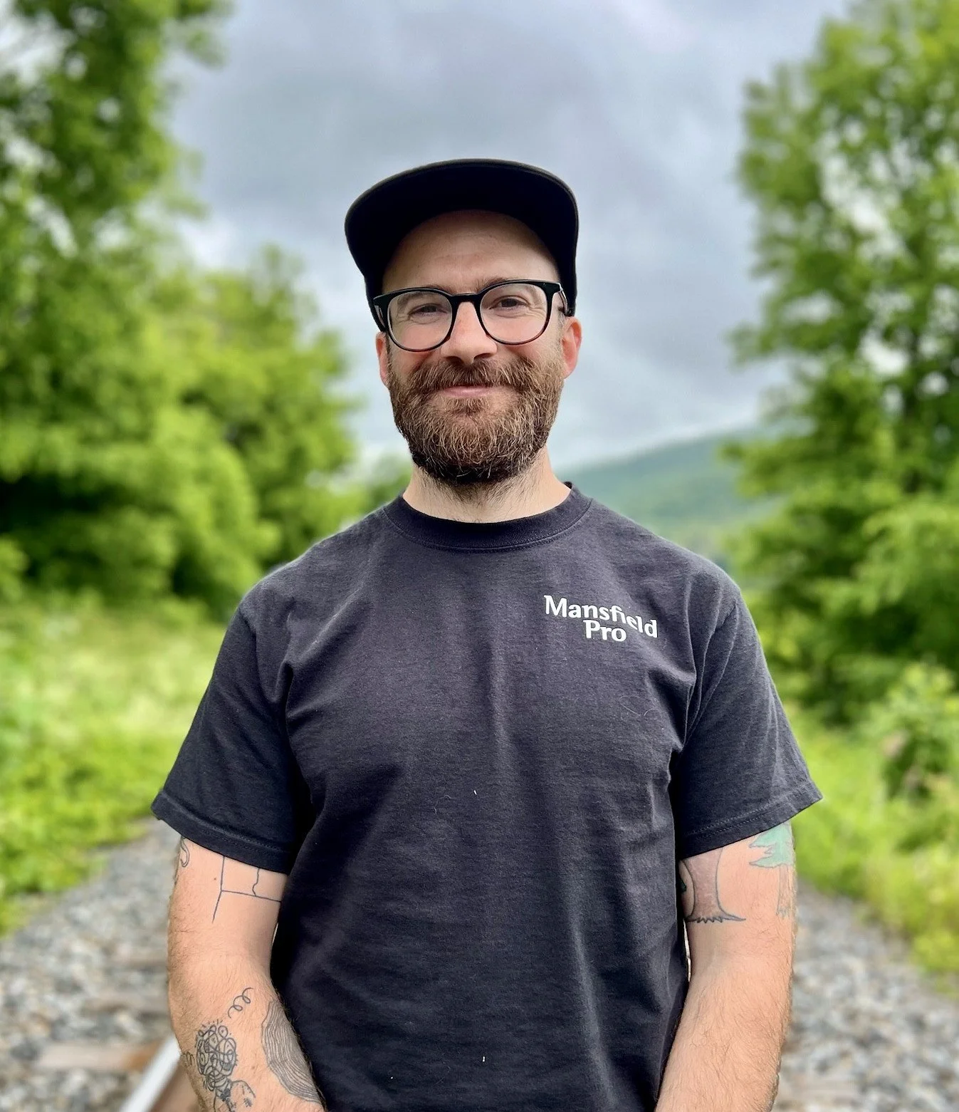 A man with glasses, a beard, and tattoos on his arms is standing outdoors on a gravel path, with green trees and a cloudy sky in the background. He is wearing a black t-shirt with the words 'Mansfield Pro' and a black cap.