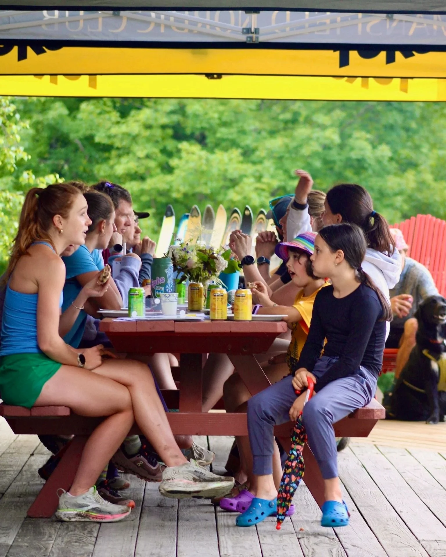 Group of children and teenagers sitting at a picnic table outdoors, engaging in conversation, with drinks and snacks on the table, surrounded by trees and colorful Adirondack chairs.