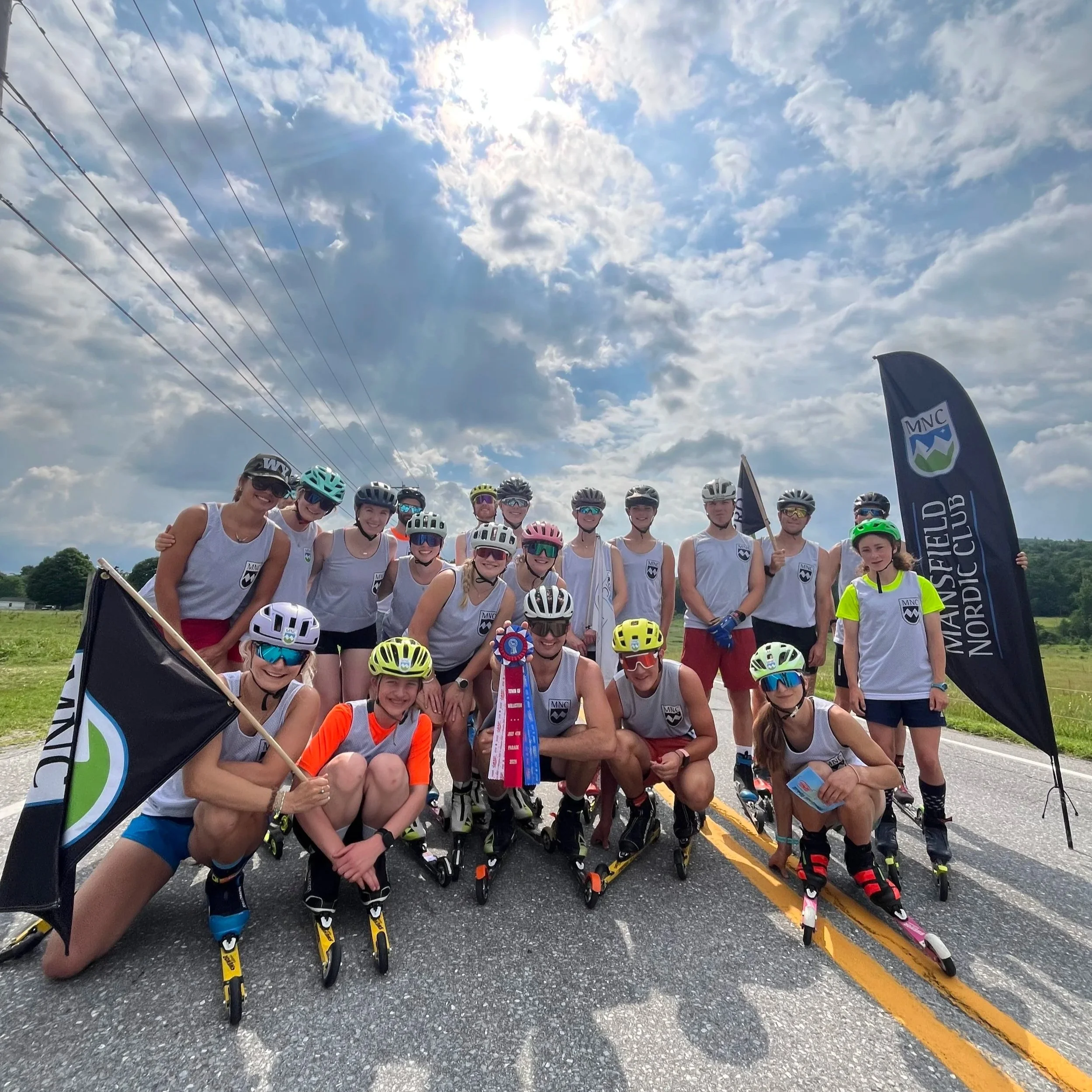Group of people dressed in rollerblades and helmets, posing together outdoors on a sunny day, with flags and a cloudy sky visible in the background.