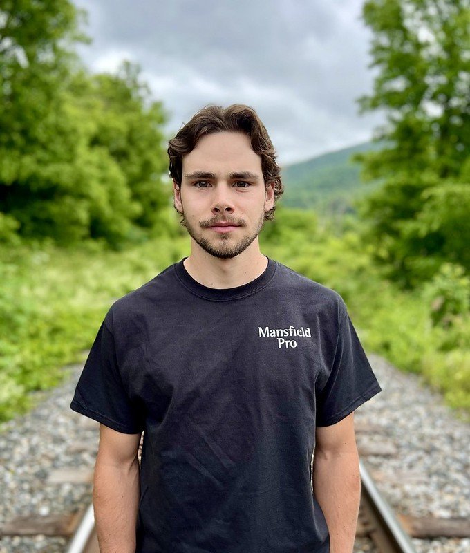 A young man with brown hair and a beard standing outdoors on a railway track, wearing a black Mansfield Pro t-shirt, with green trees and a cloudy sky in the background.