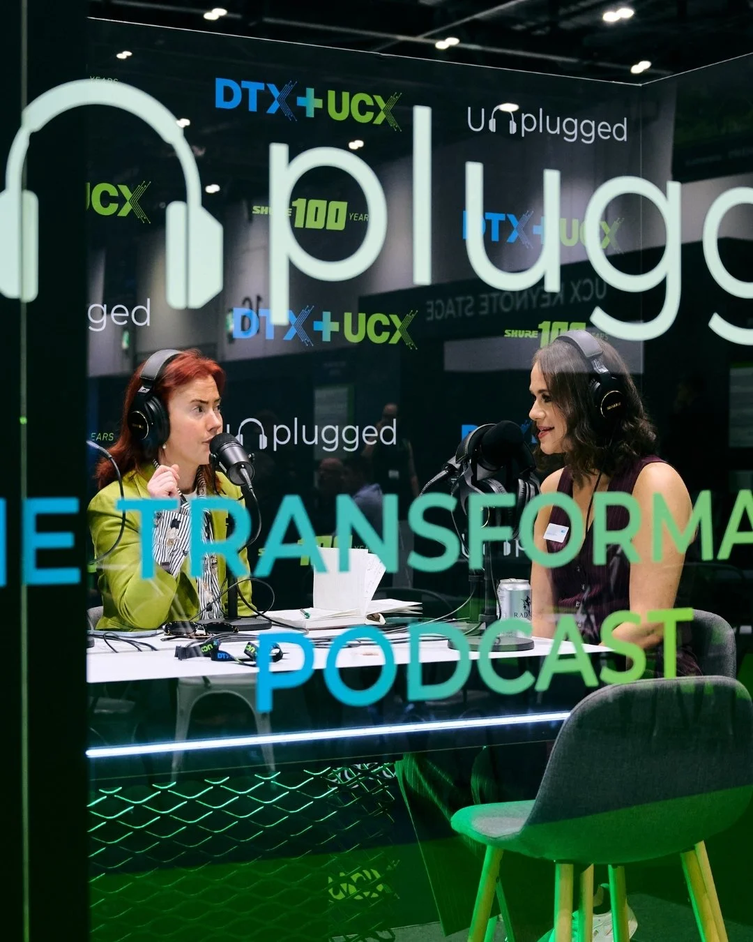 Two women with headphones recording a podcast in a studio with glass window, decorated with colorful words and logos, including 'The Transformative Podcast' and 'DTX+UCX'.