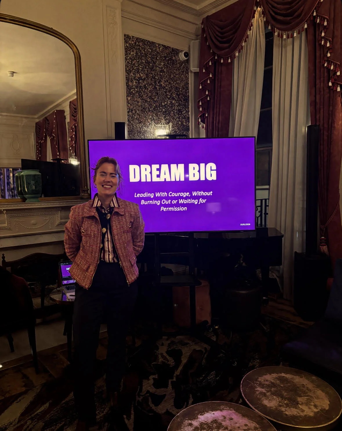 Woman smiling and standing in front of a large purple screen with the words 'DREAM BIG' and a quote, in a decorated room with draped curtains and a mirror.