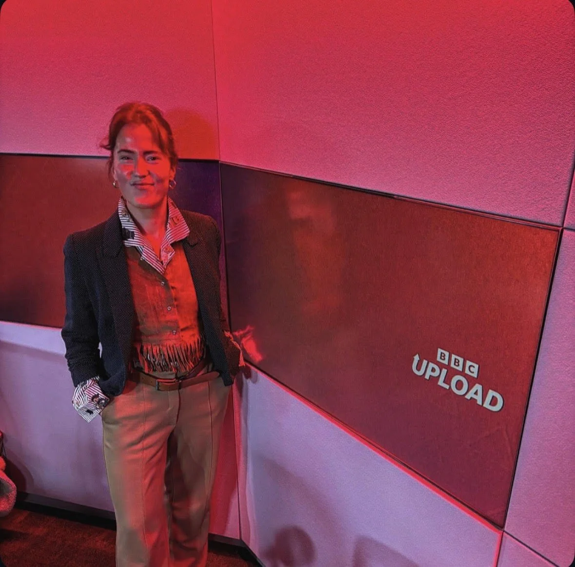A woman standing in front of a red and pink wall with a BBC ULOAD sign, wearing a blazer, striped shirt, and beige pants.