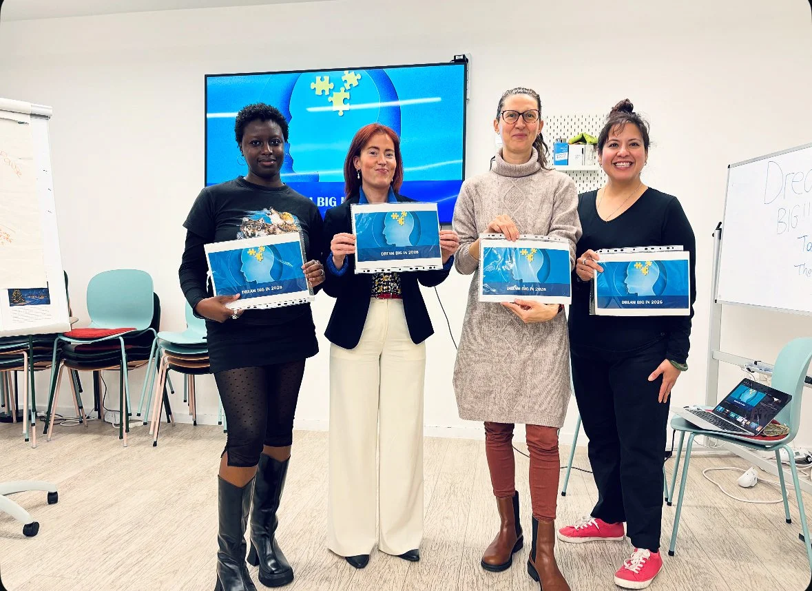 Four women standing in a room, each holding a calendar with a blue cover featuring a globe and puzzle piece design. Behind them, a large screen displays a similar image. To the left, a whiteboard and stack of chairs are visible, and on the right, a small table with a laptop and some scattered items.