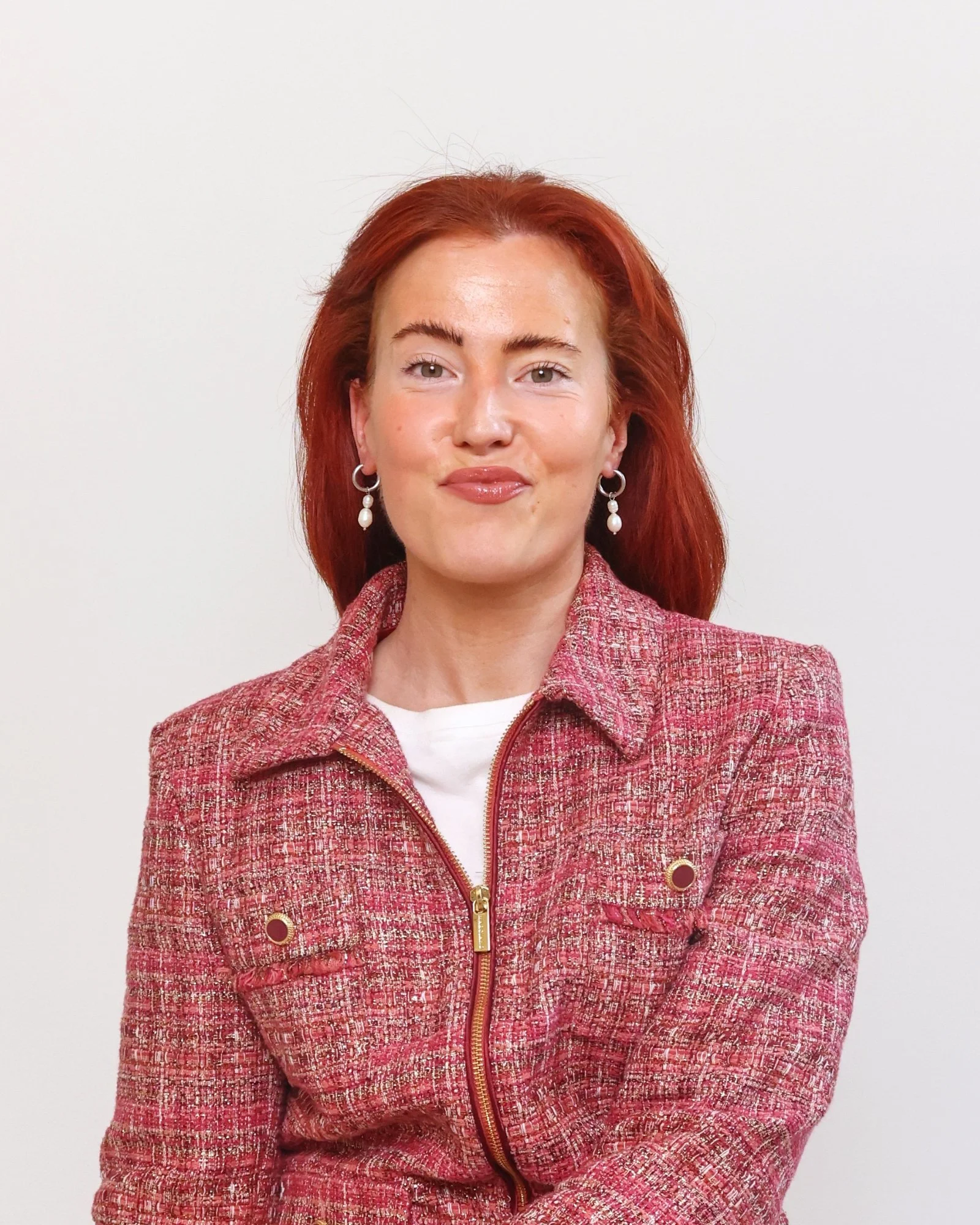 A woman with red hair, wearing pearl earrings and a pink tweed jacket, standing against a plain white background.