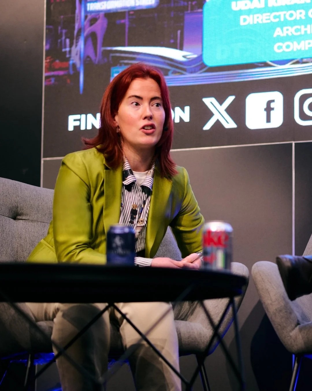A woman with reddish-brown hair wearing a green jacket and striped blouse speaking at a panel or event with a digital screen behind her displaying various icons and text.