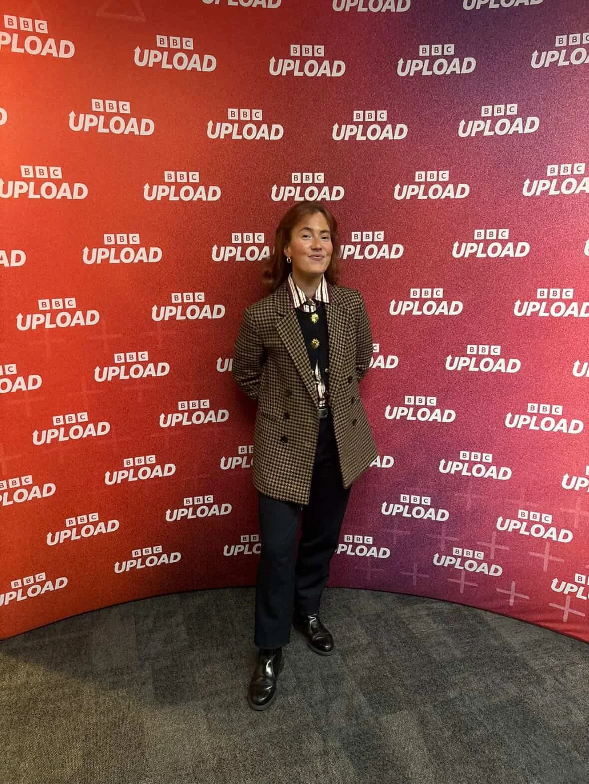 A woman standing in front of a red background with multiple 'BBC UPLOAD' logos, wearing a checkered blazer, striped shirt, black pants, and black boots.