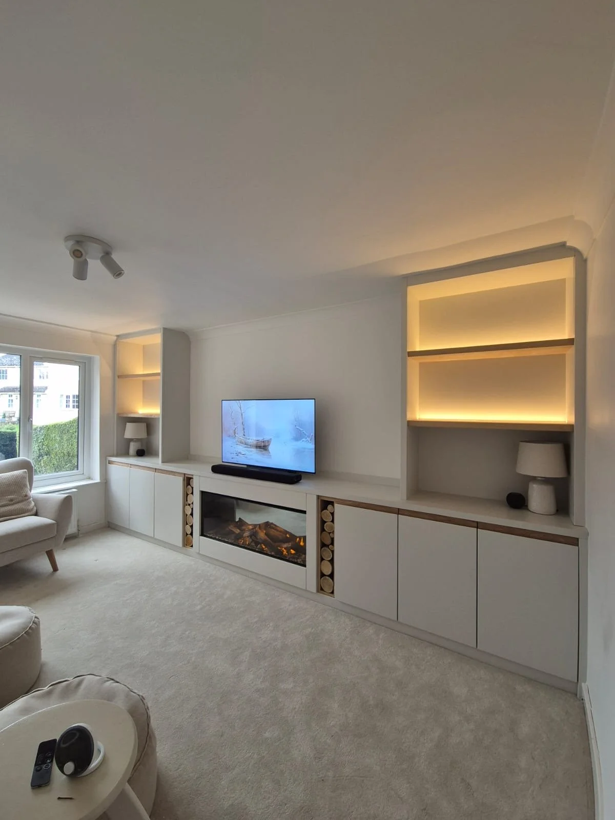 Living room with beige carpet, white furniture, a window, a TV on a white entertainment unit, some built-in shelves with decor, one appearing to be a fireplace, and a small round side table with remote and speaker.