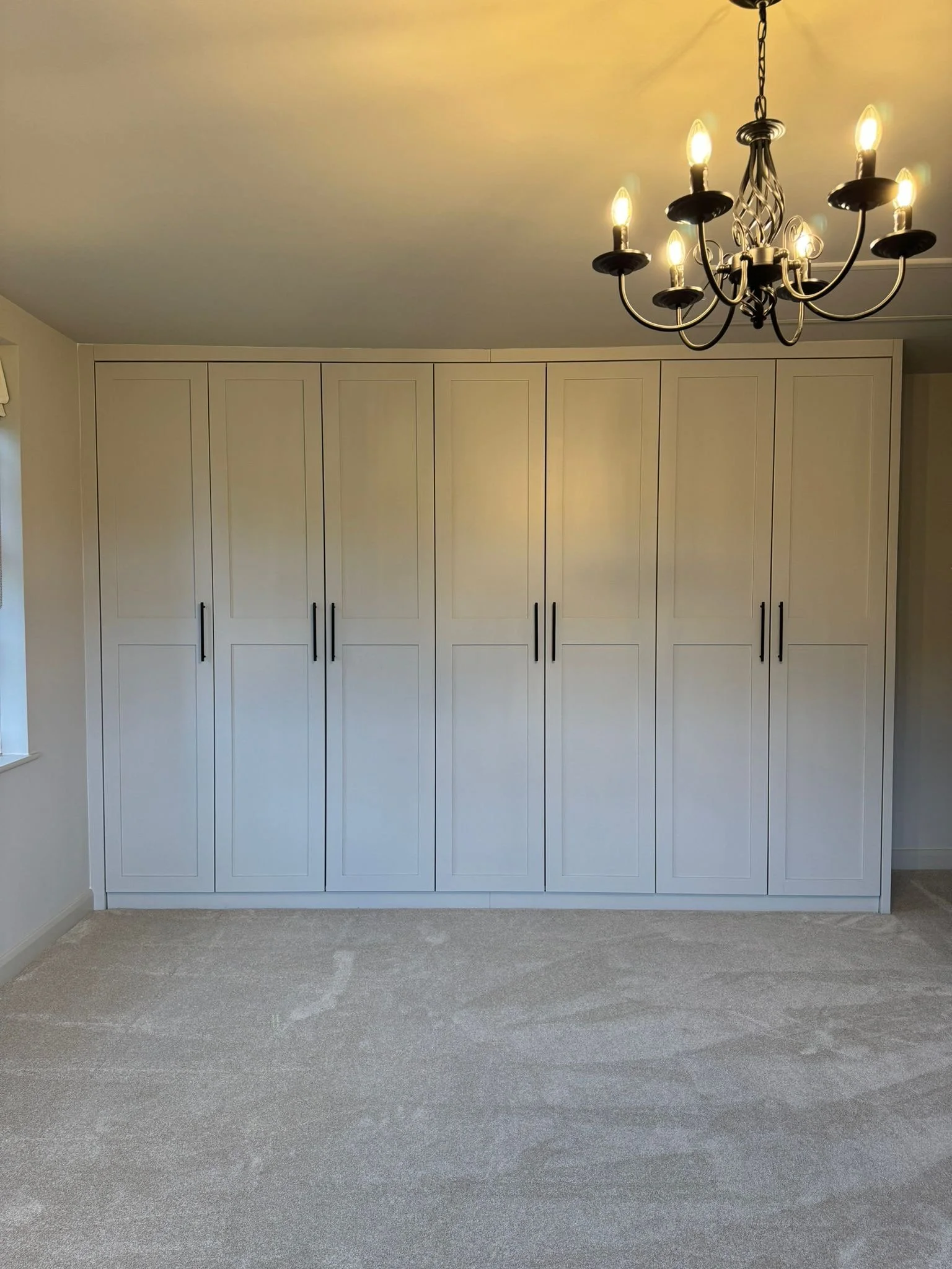 Room with a large, closed white wardrobe, a chandelier with multiple lights, a small part of a window on the left, and a beige carpeted floor.