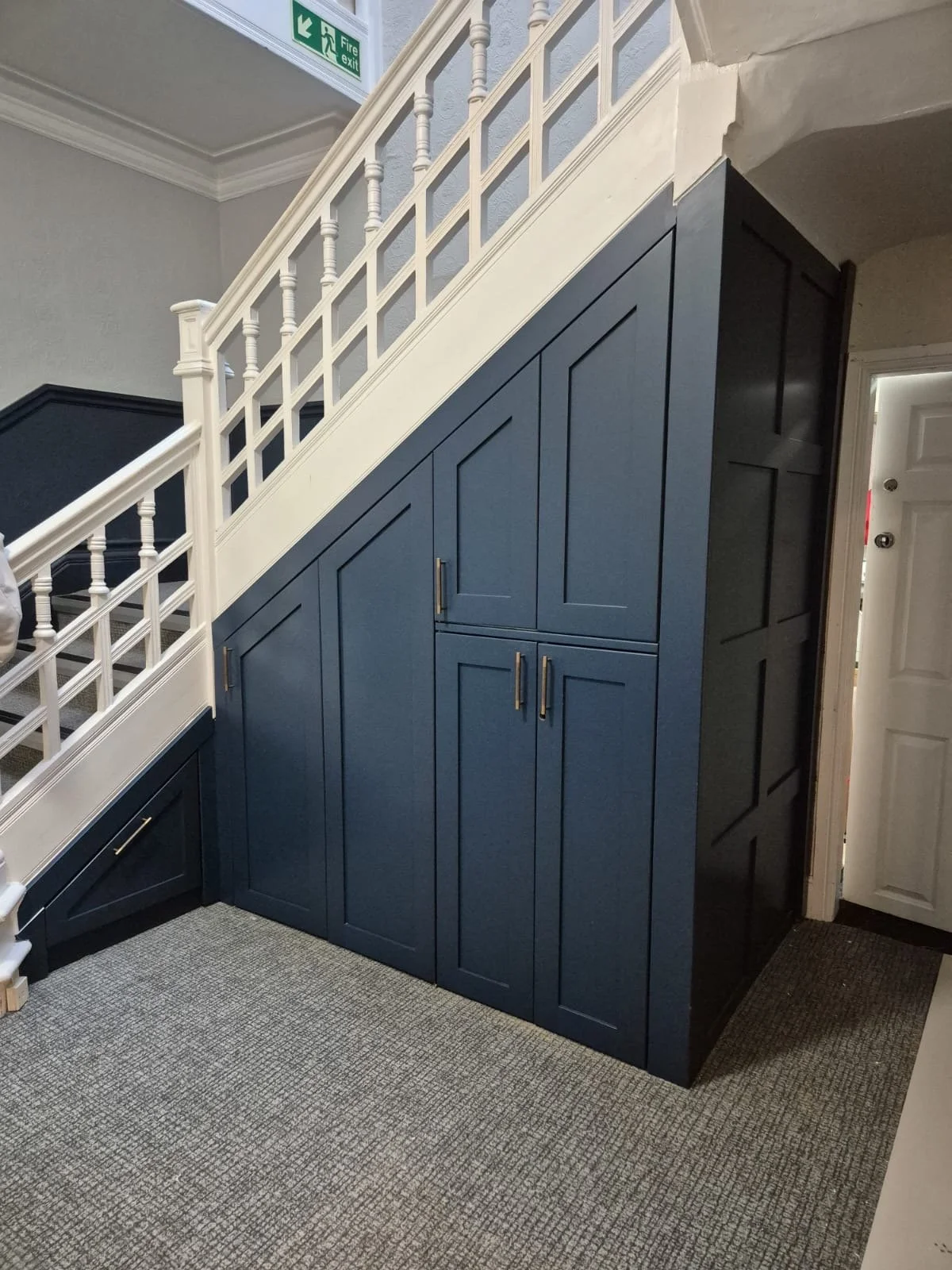 A built-in navy blue storage cabinet underneath a staircase with white railing and spindles, located in a home interior. A door is visible to the right.