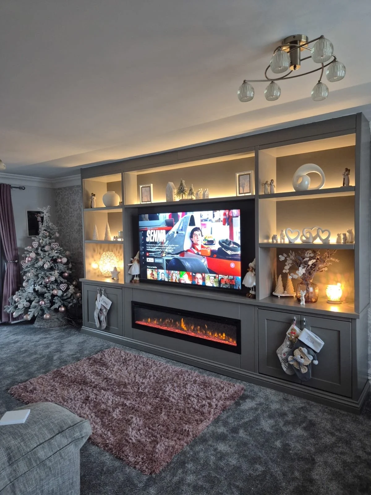 Living room featuring a gray entertainment center with a TV, electric fireplace, Christmas tree, and holiday decorations.