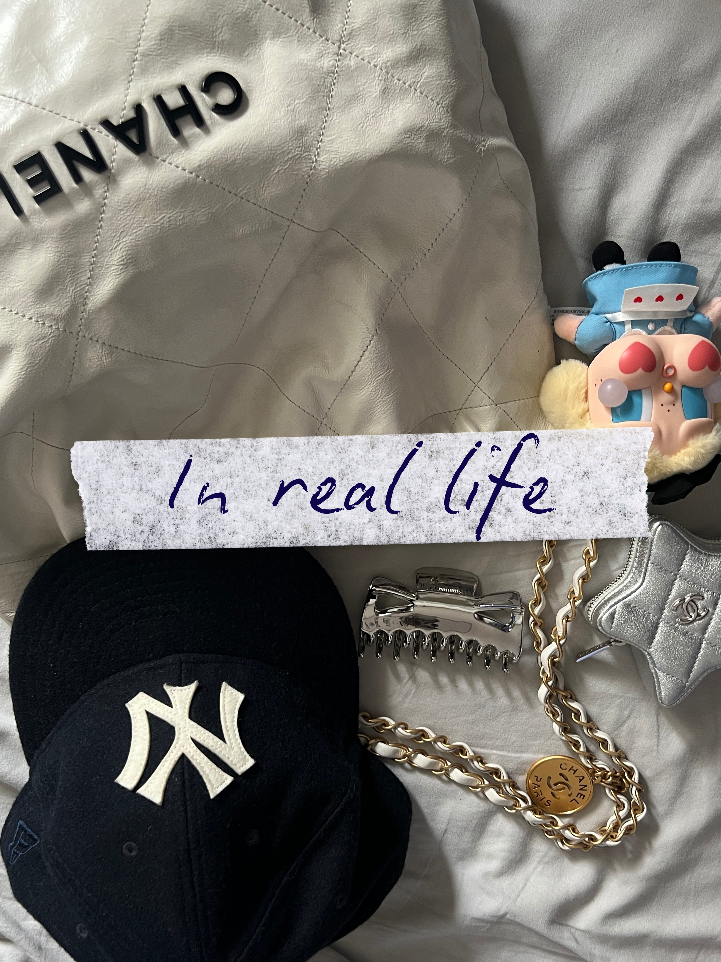 Une casquette de baseball noire avec le logo NY, une pince à cheveux argentée, une chaîne en or avec un pendentif, une petite sacoche en cuir argentée, une poupée en peluche avec un costume de pompier, et un jouet en forme de personnage avec un visag
