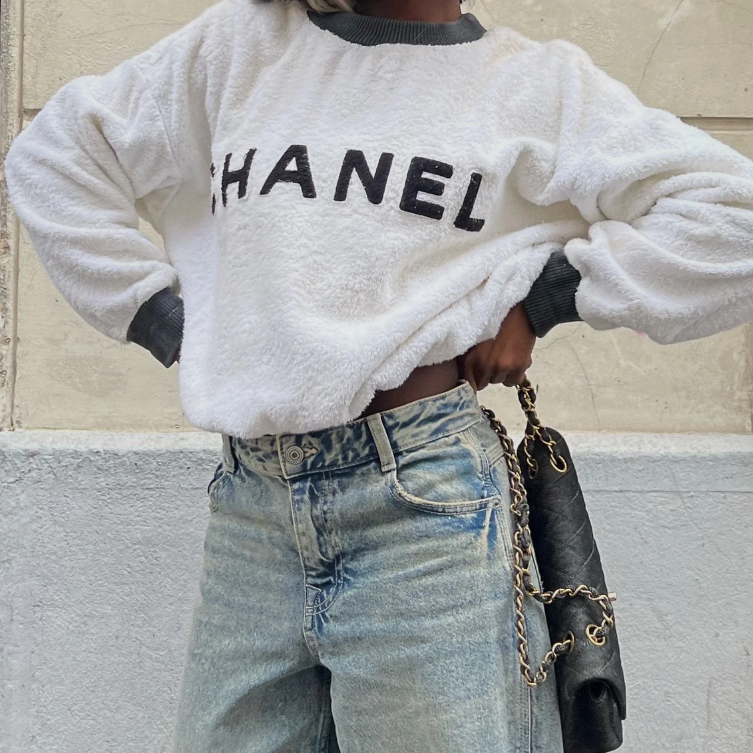Une personne portant un sweat blanc en peluche avec le mot "CHANEL" en lettres noires, des jeans décontractés, et tenant un sac à main noir avec une chaîne dorée, devant un mur beige.