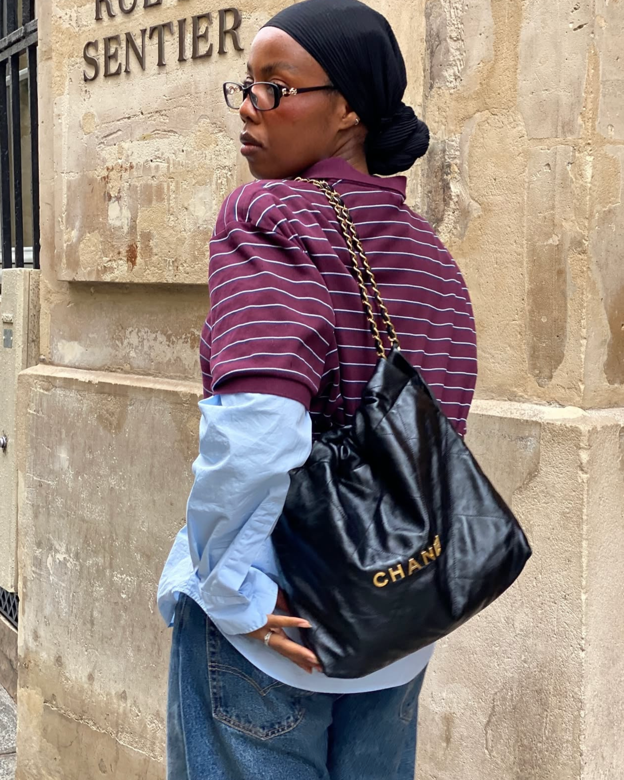 Femme portant un sac à main noir de la marque Chanel. Elle porte une chemise à manches longues et un t-shirt rayé bordeaux et blanc, ainsi qu'un foulard noir sur la tête. Elle se tient contre un mur en pierre avec une inscription en français.
