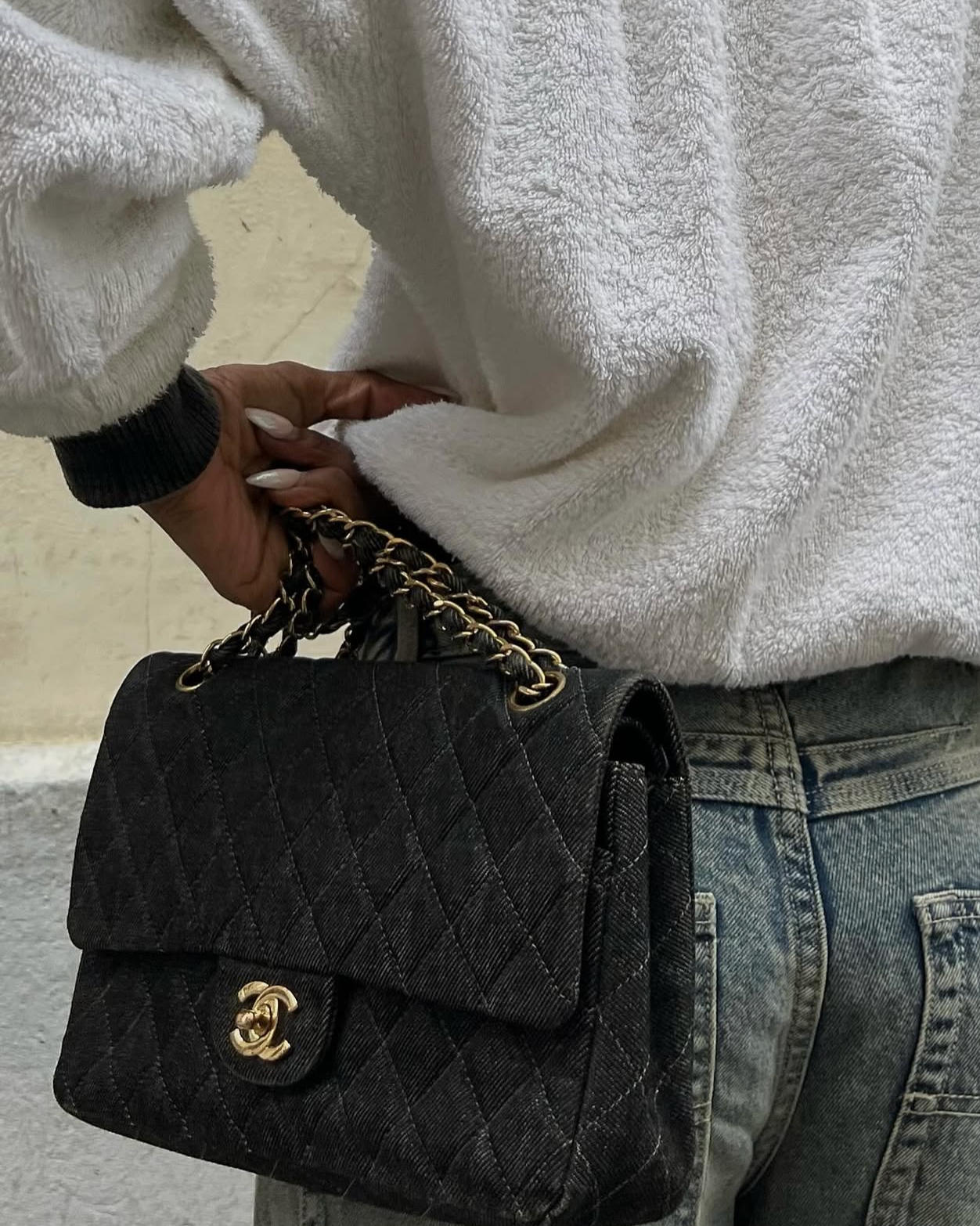 Personne tenant un sac à main noir en tweed avec chaîne dorée, portant un pull blanc en molleton et un jean bleu, près d'un mur beige.