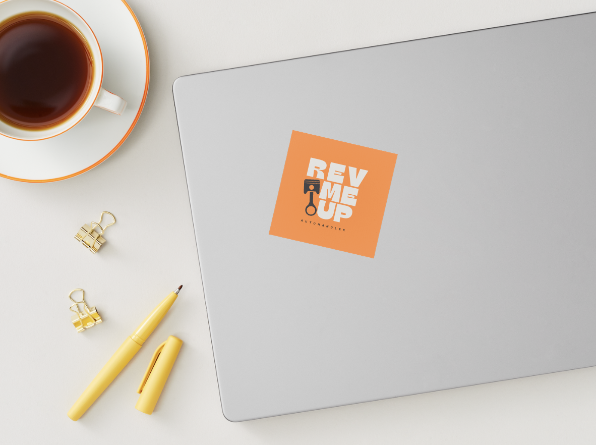 A closed laptop with a sticker logo that says "REV ME UP" with a graphic of a car key, on a white desk with a cup of coffee on a saucer, yellow pen, and two gold binder clips.