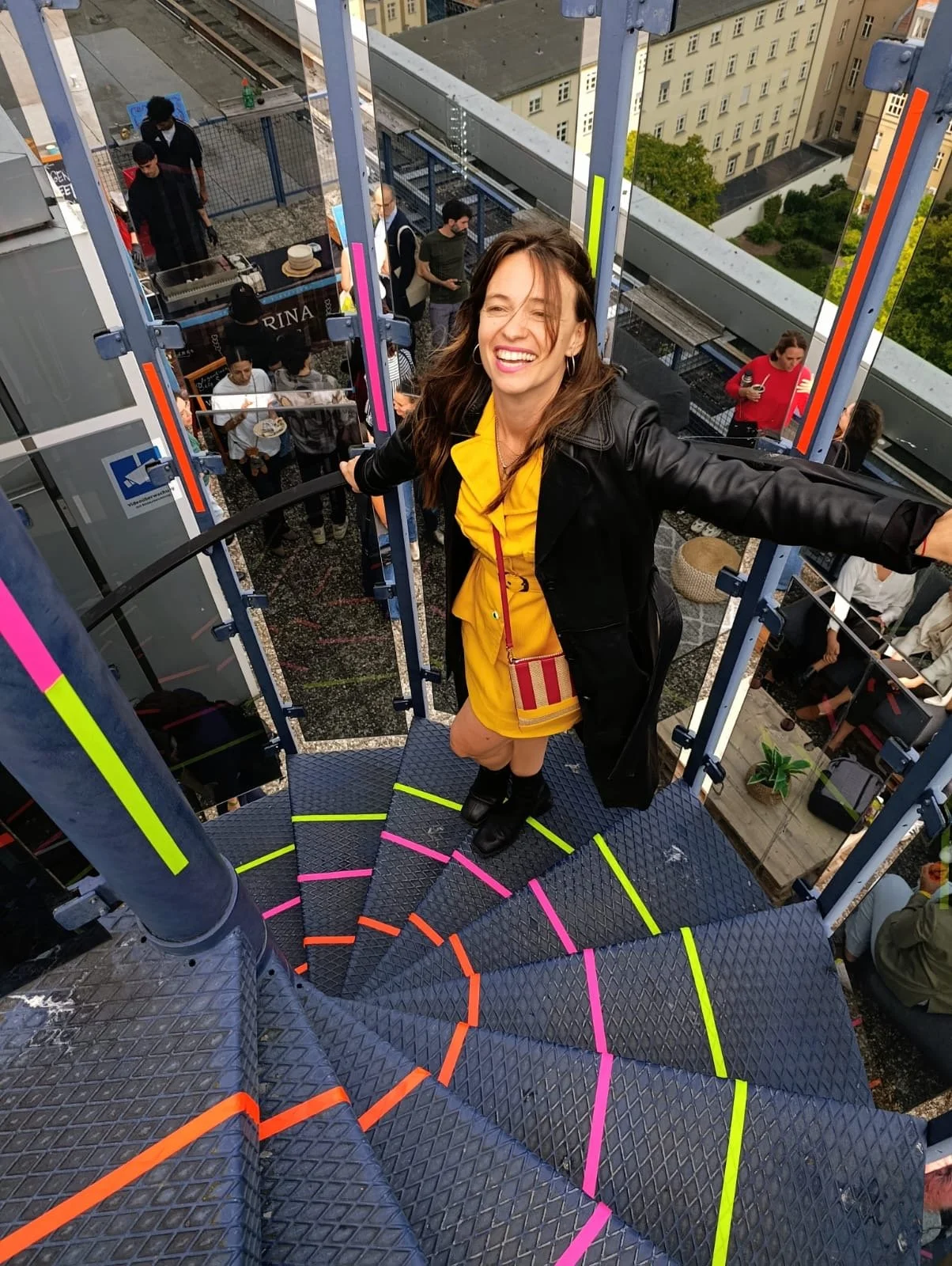 Dana Schröck, founder of De Nada creative studio,  in a yellow dress and black jacket smiling and posing on a spiral staircase on a rooftop with her art intervention, with people in the background attending a social event.
