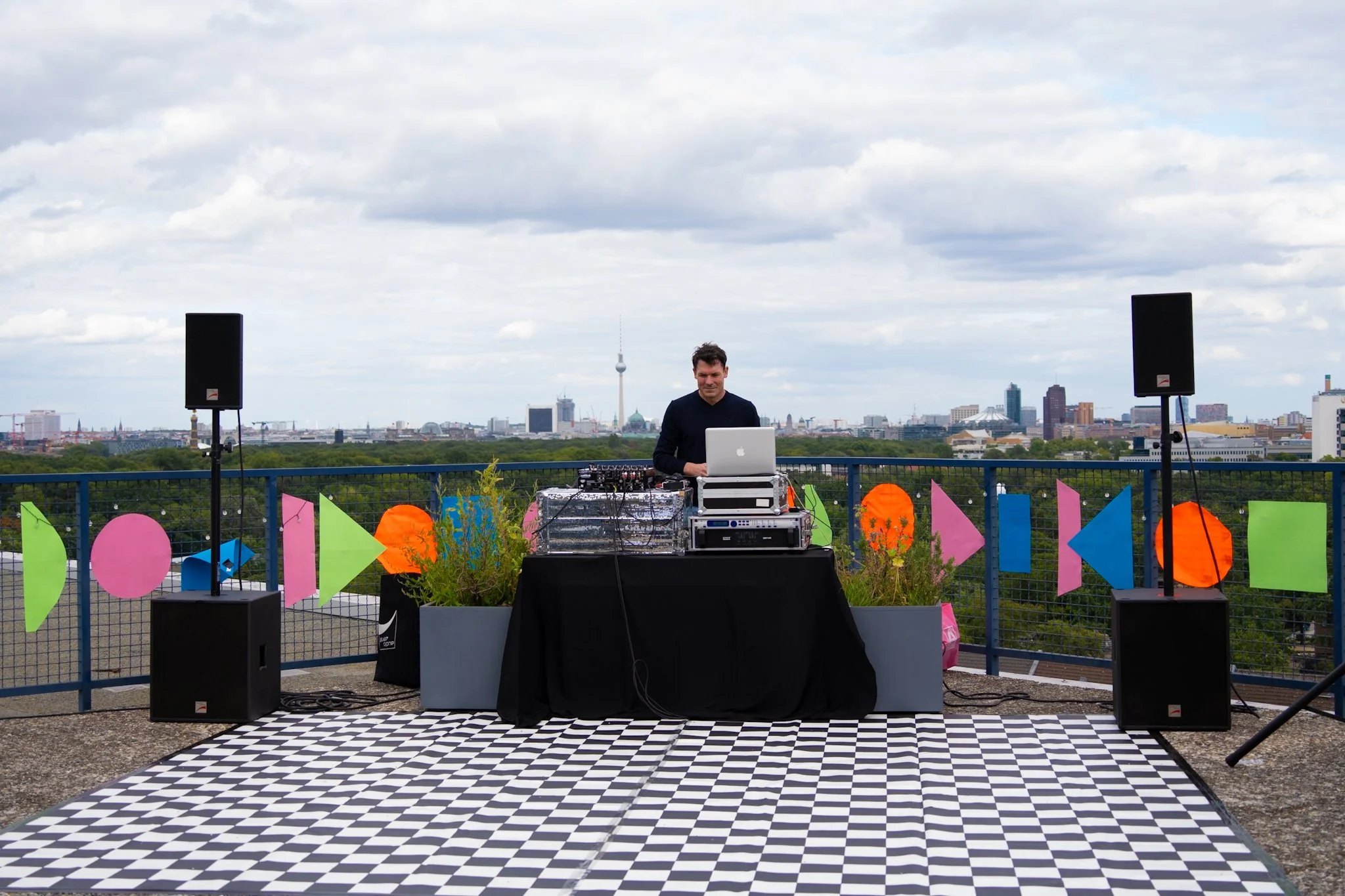 Event decoration: DJ performing on an outdoor rooftop with a city skyline in the background, colorful geometric decorations on the railing, and a checkered dance floor in front.