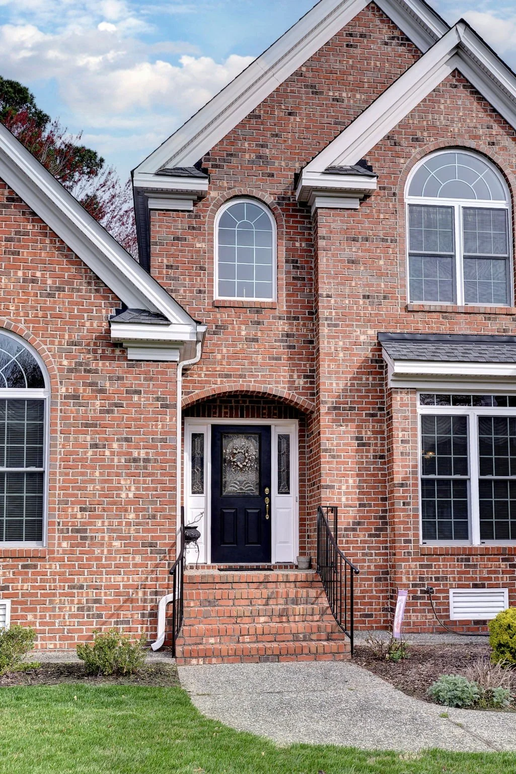 Front entrance of a brick house with a black door and steps, leading to a sidewalk and lawn in front.