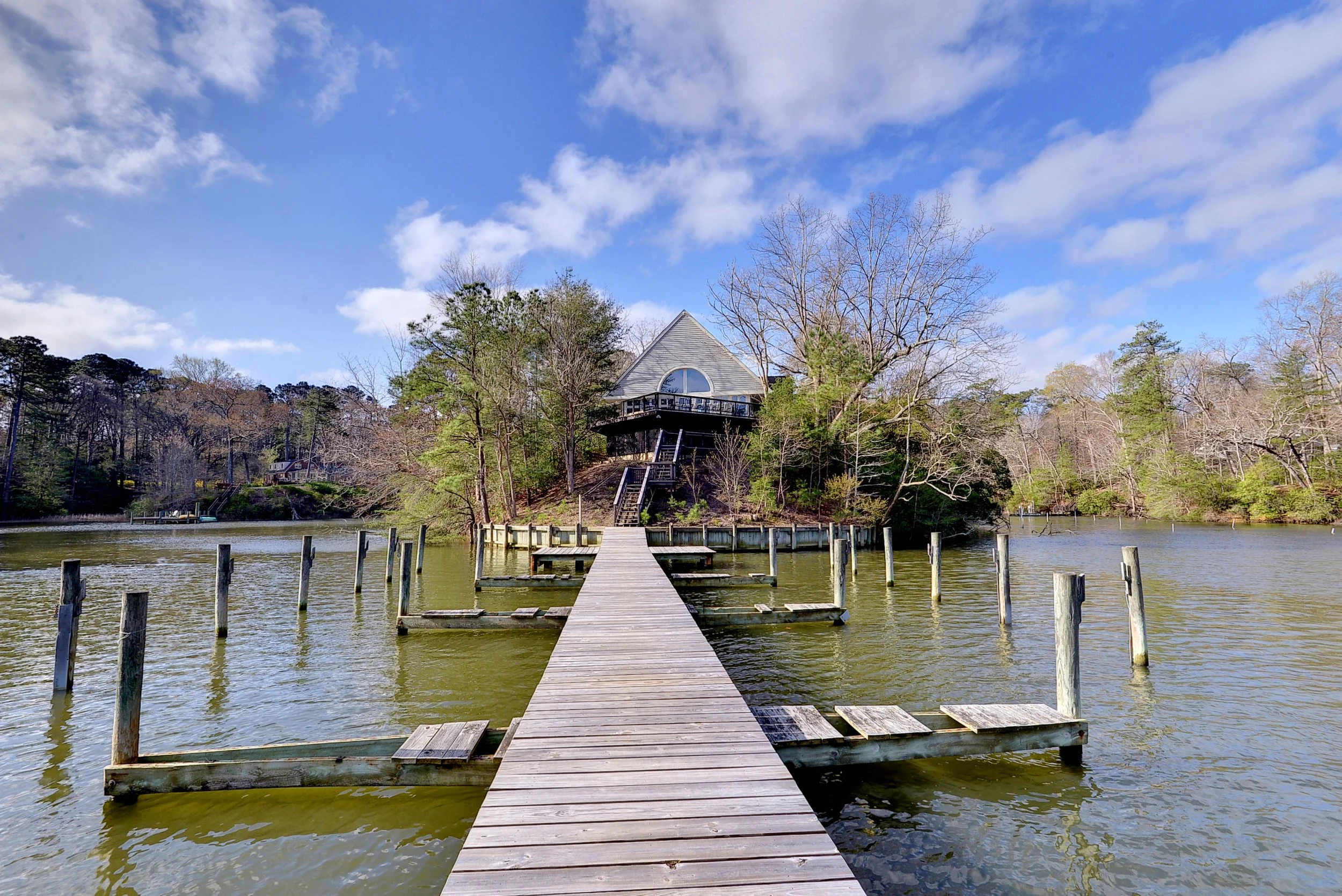 A wooden dock extends over a lake leading to a house on a hill surrounded by trees, with a partly cloudy sky above.
