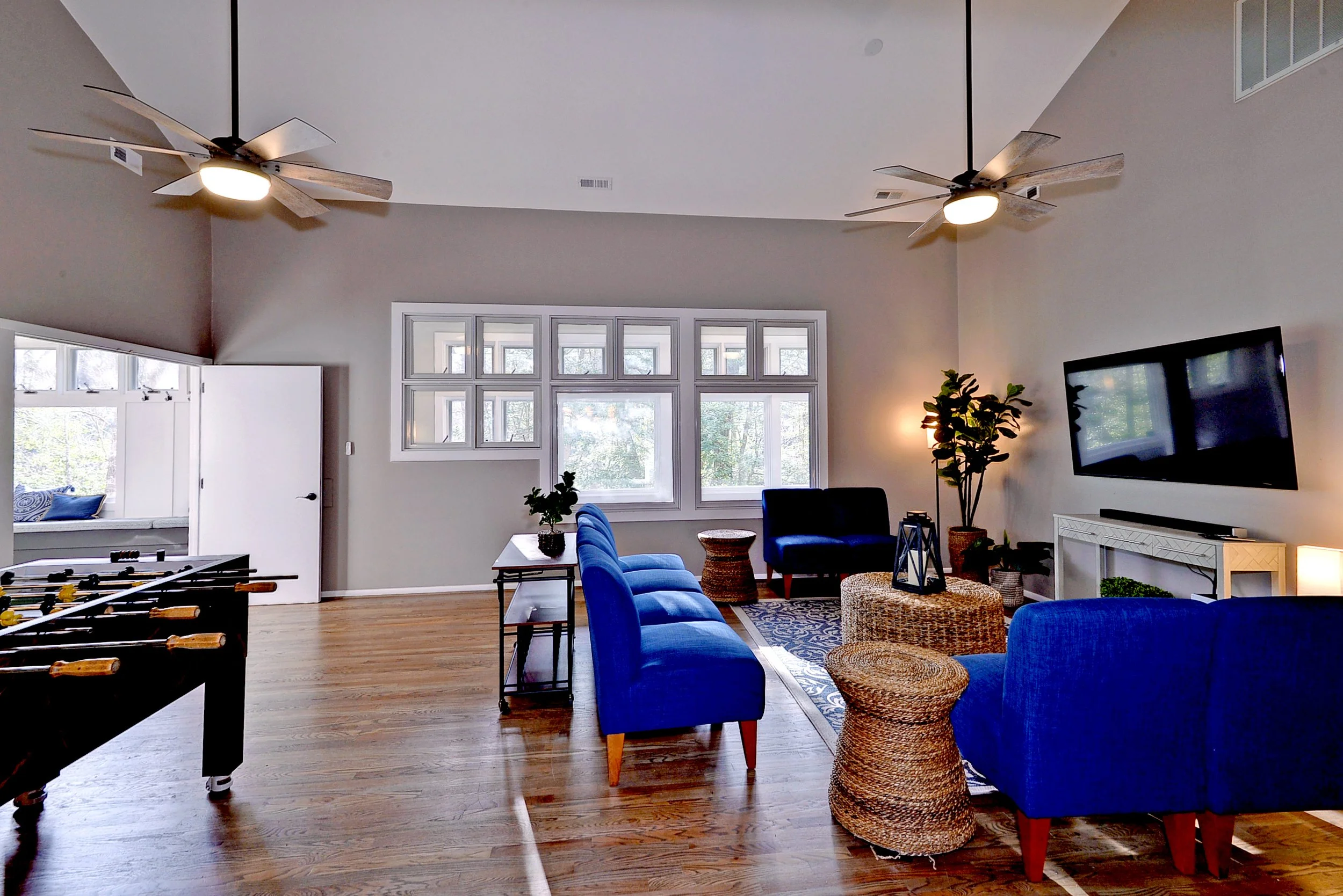 Living room with hardwood floors, blue sofas, a wall-mounted TV, and large windows. There's a foosball table on the left and potted plants around the room.