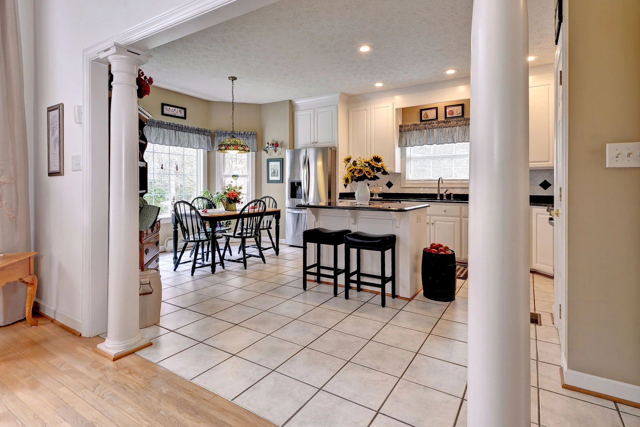 View of a bright, open kitchen and dining area with tiled floor, white cabinetry, black countertops, island with two black barstools, and a dining table with six black chairs. Sunlight streams through multiple windows with checkered valances. Decor i