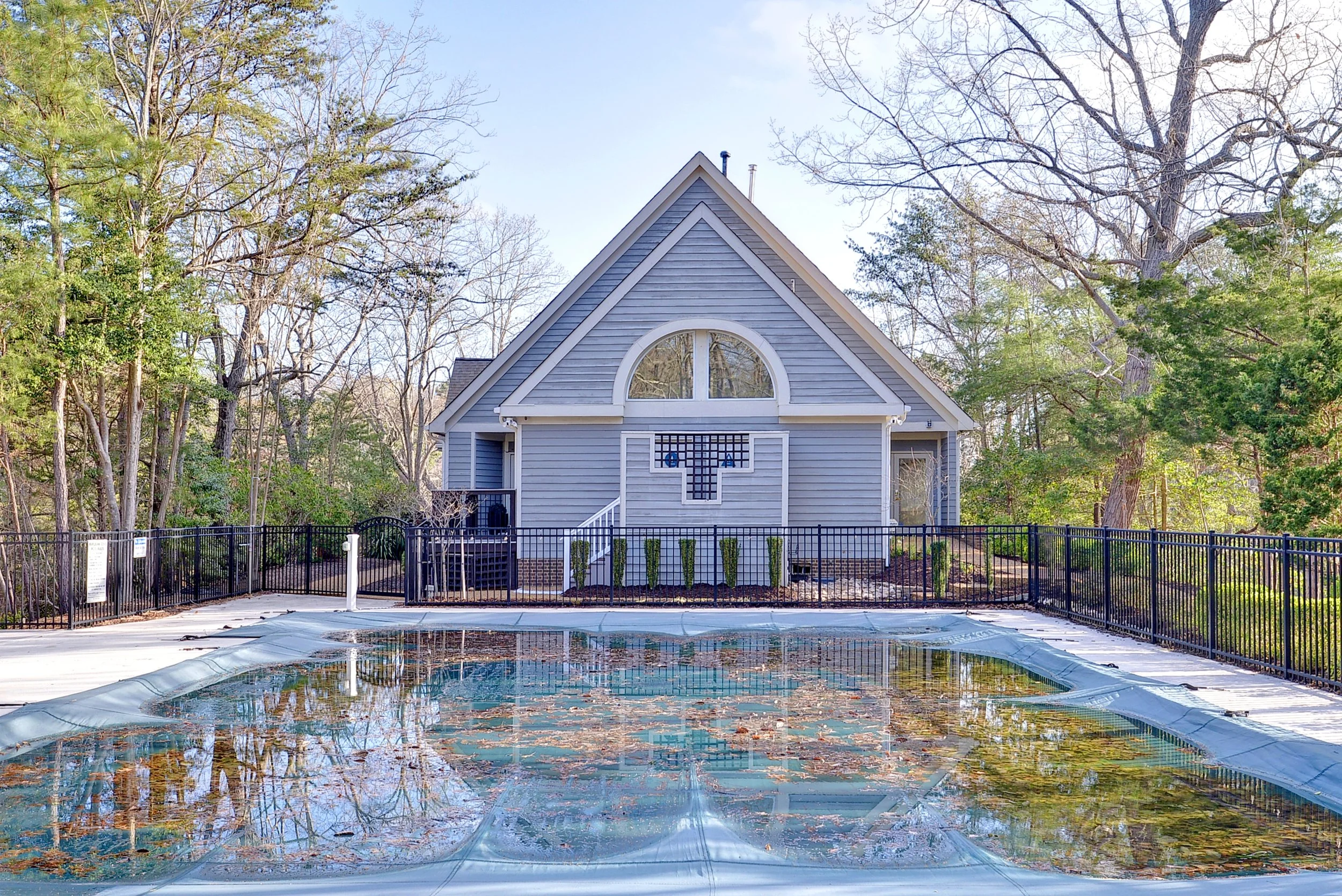 A gray house with a steep roof, surrounded by trees, with a fenced backyard containing a swimming pool covered with fallen leaves.